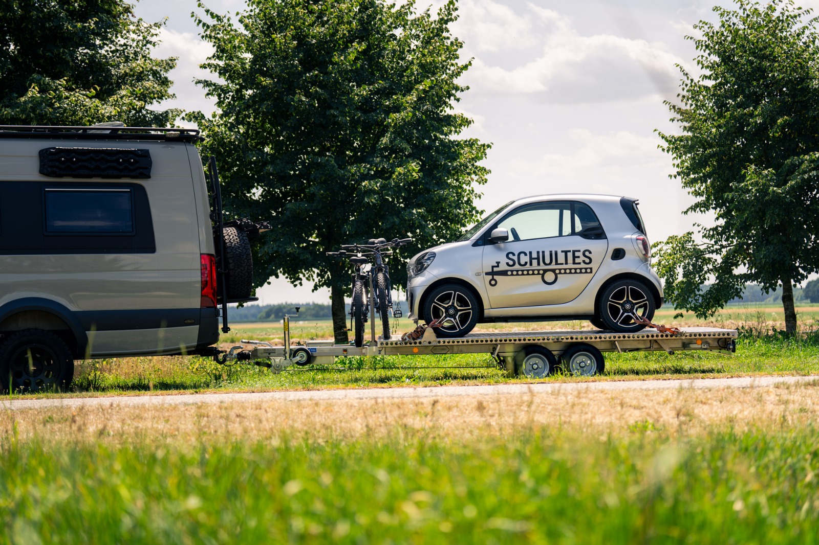 UA350 Tandem Anhänger Smart und Fahrräder Anhänger aus Aluminium mit Smart fortwo und zwei Fahrrädern hinter einem Mercedes Sprinter vor Bäumen und einer grünen Wiese