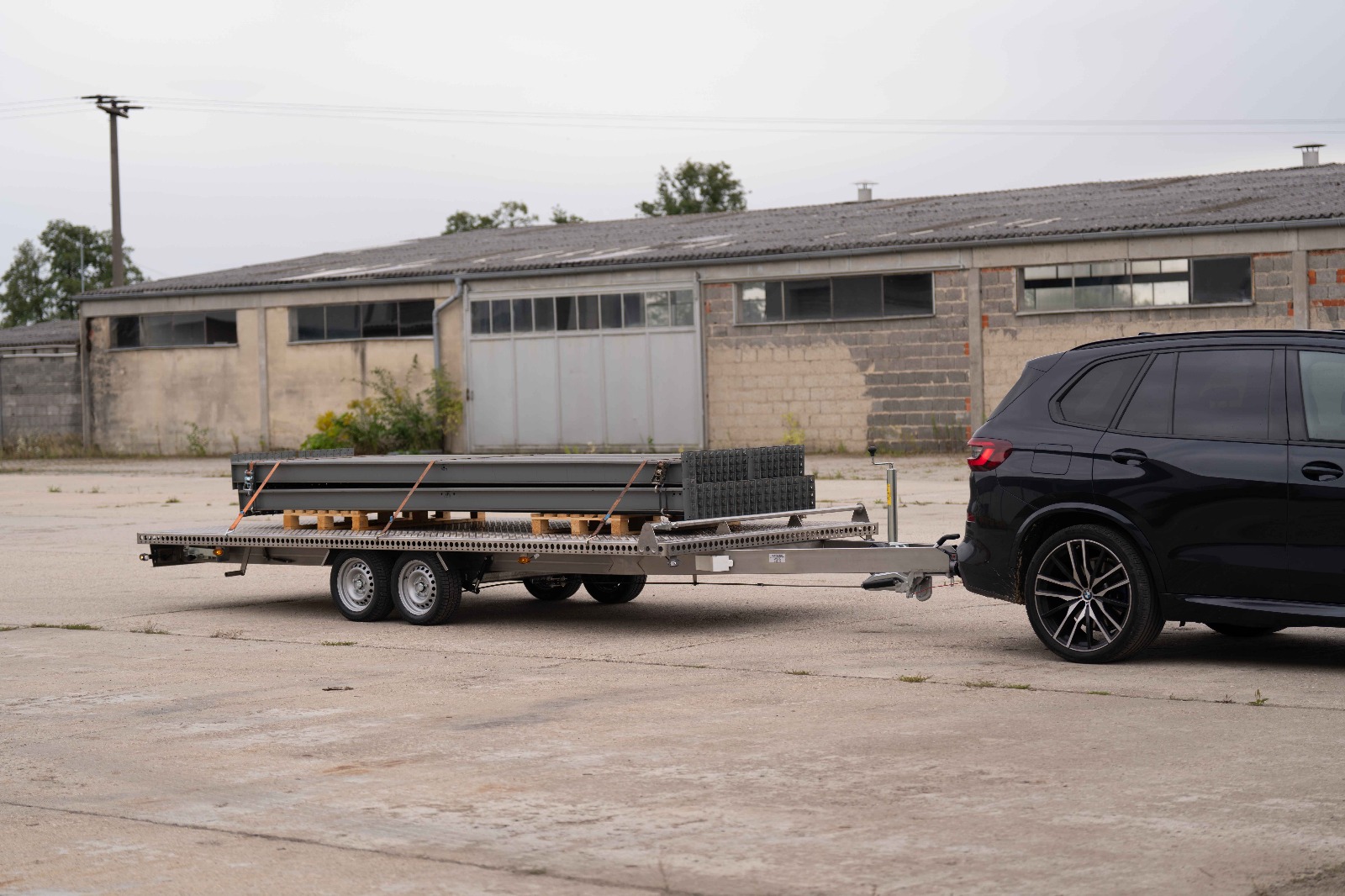 UA510 Edelstahl Plattform mit Regalträger 2 Edelstahl Plattformanhänger hinter BMW X5. Anhänger ist mit Regaltraversen beladen vor einem alten Industriegebäude