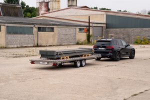 Edelstahl Plattformanhänger hinter BMW X5. Anhänger ist mit Regaltraversen beladen vor einem alten Industriegebäude