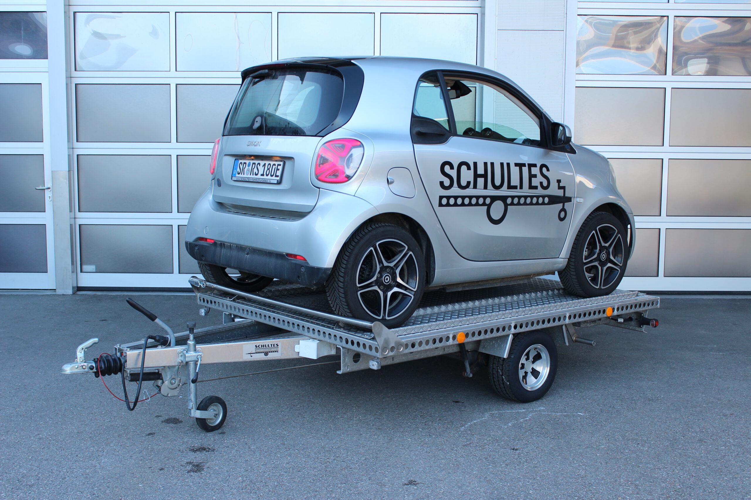 UA250 Universalanhänger aus Aluminium mit Zubehör für den Transport eines Smart fortwo. Smart fortwo auf dem Anhänger rückwärts geladen in silber, vor einem Hallentor