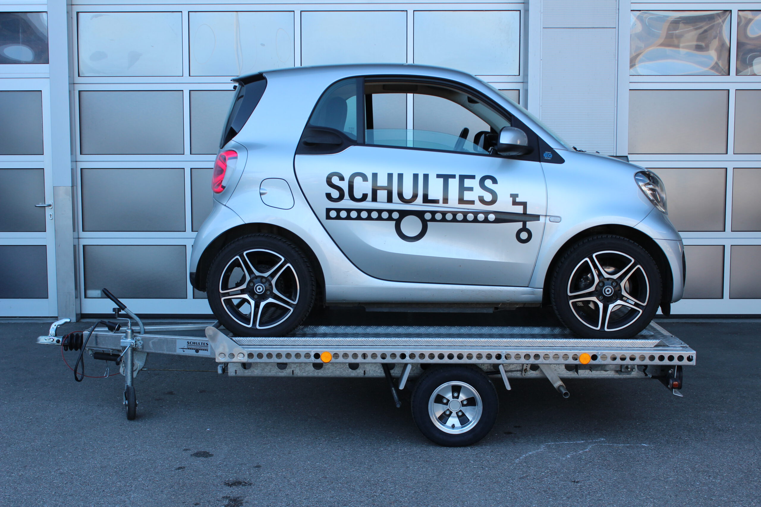 UA250 Universalanhänger aus Aluminium mit Zubehör für den Transport eines Smart fortwo. Smart fortwo auf dem Anhänger rückwärts geladen in silber, vor einem Hallentor