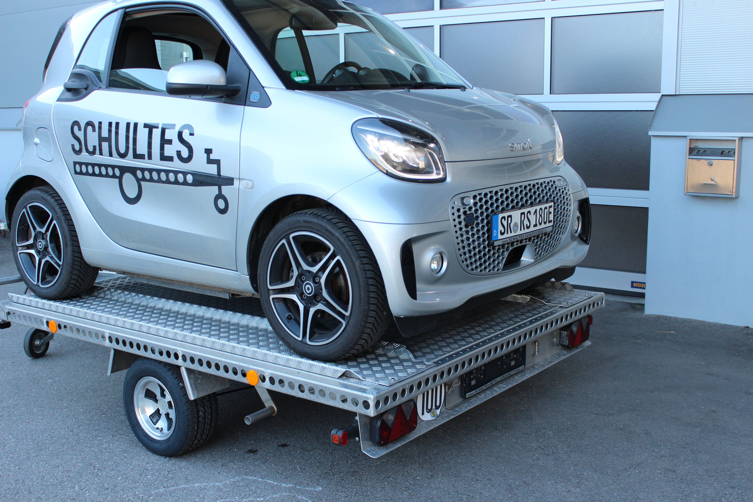 UA250 Universalanhänger aus Aluminium mit Zubehör für den Transport eines Smart fortwo. Smart fortwo auf dem Anhänger rückwärts geladen in silber, vor einem Hallentor