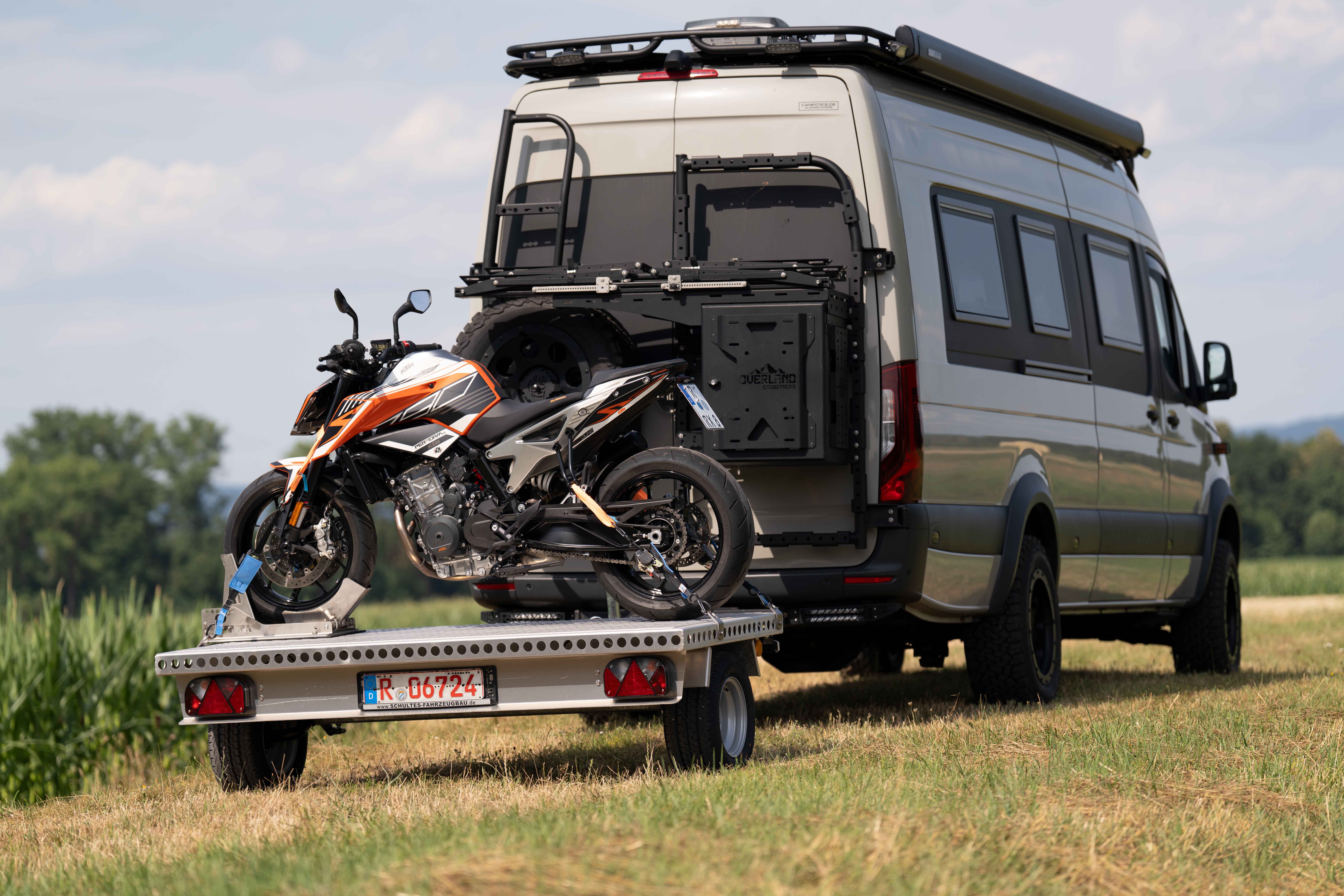 Kleiner Aluminium Plattformanhänger mit einer KTM Duke beladen hinter einem grünen Mercedes Sprinter auf einer grünen Wiese