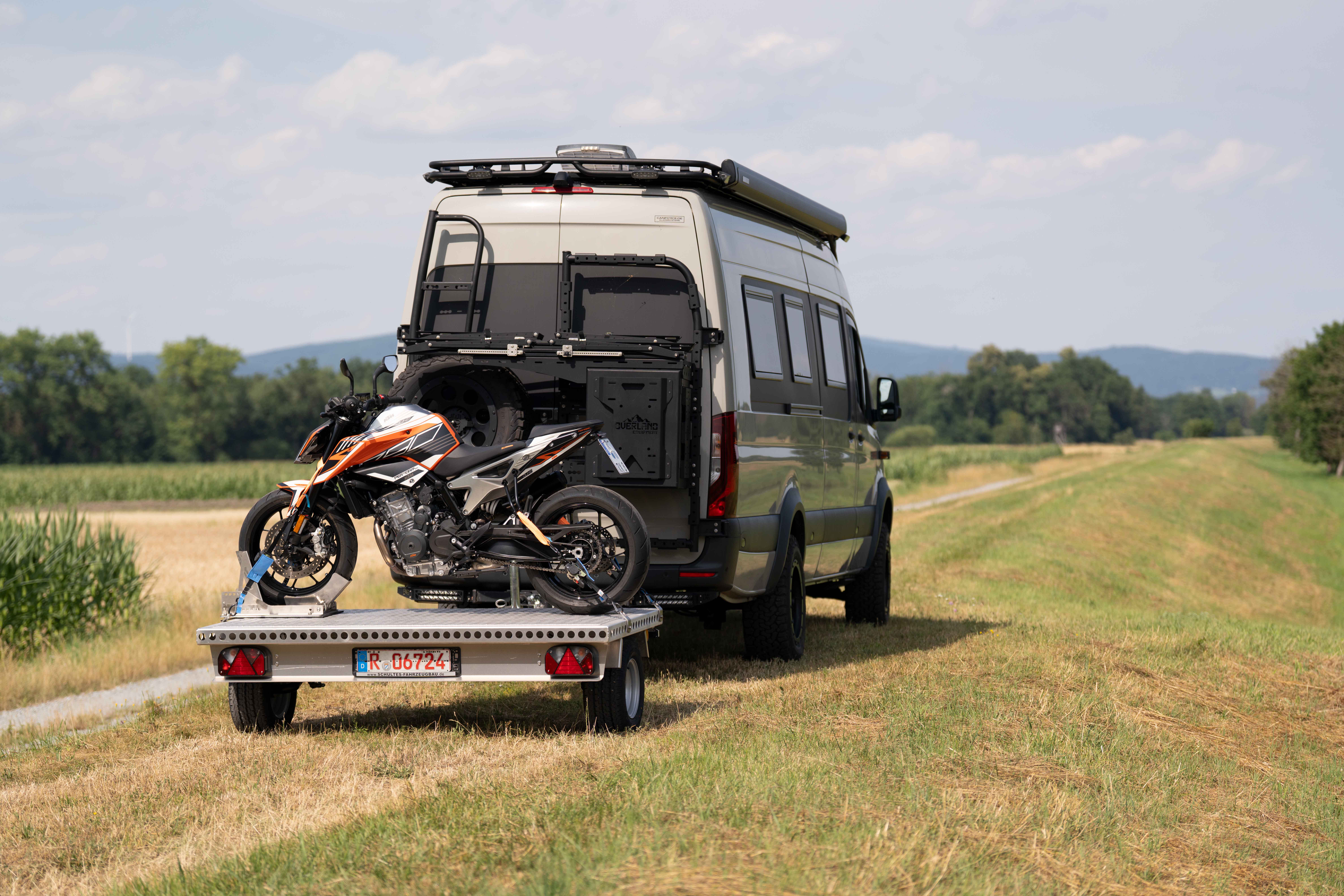 Kleiner Aluminium Plattformanhänger mit einer KTM Duke beladen hinter einem grünen Mercedes Sprinter auf einer grünen Wiese
