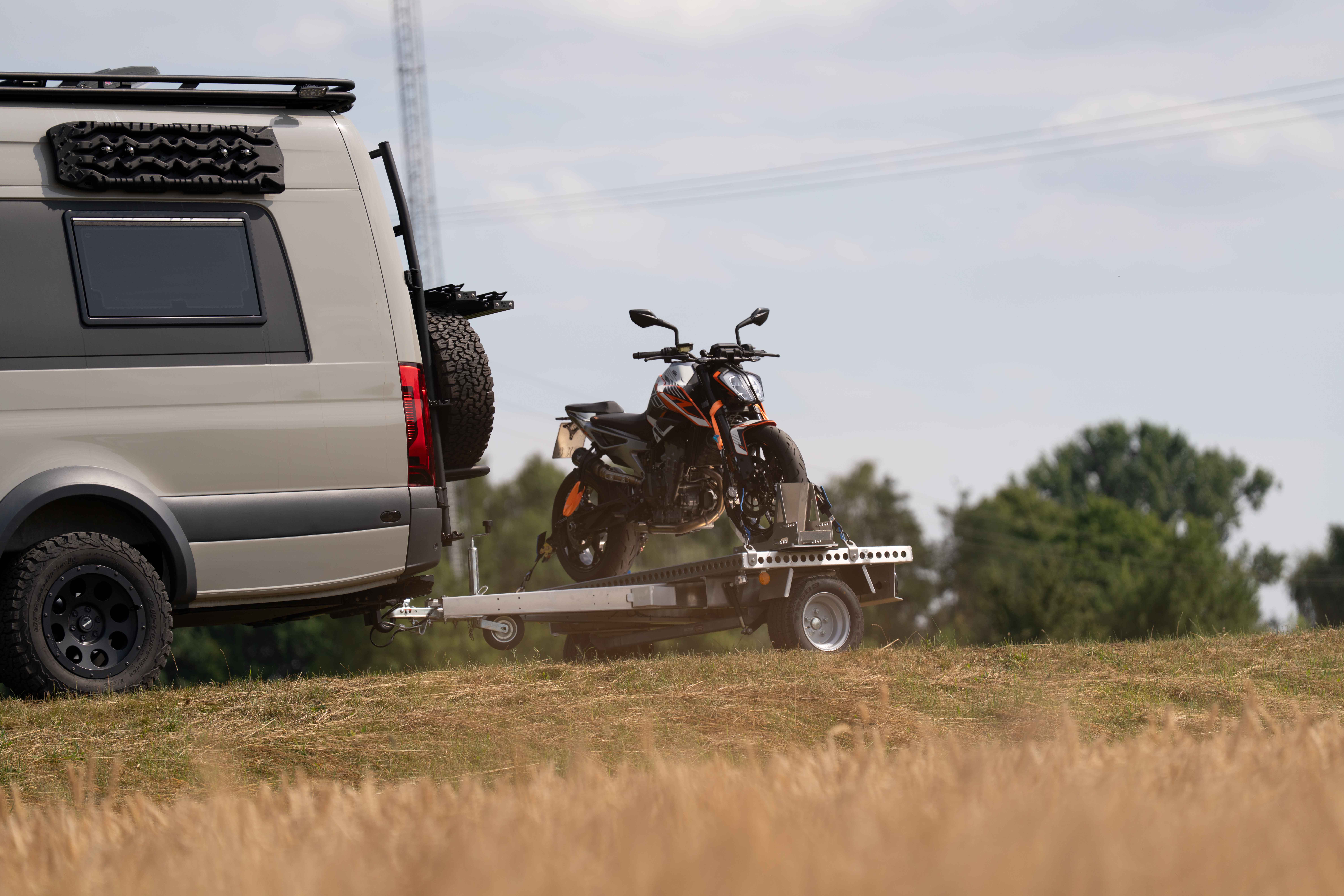 Kleiner Aluminium Plattformanhänger mit einer KTM Duke beladen hinter einem grünen Mercedes Sprinter auf einer grünen Wiese
