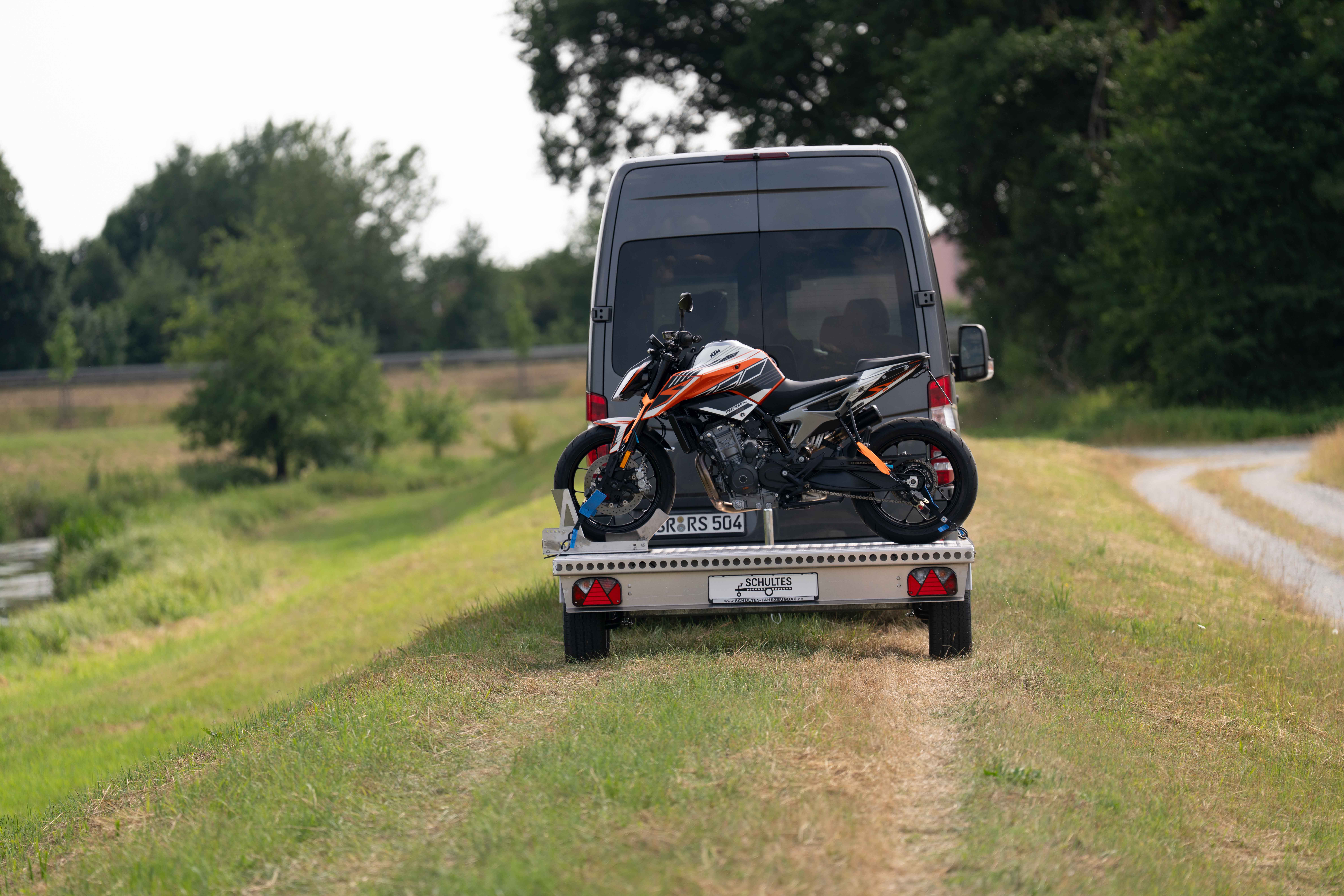 Kleiner Aluminium Plattformanhänger mit einer KTM Duke beladen hinter einem grünen Mercedes Sprinter auf einer grünen Wiese