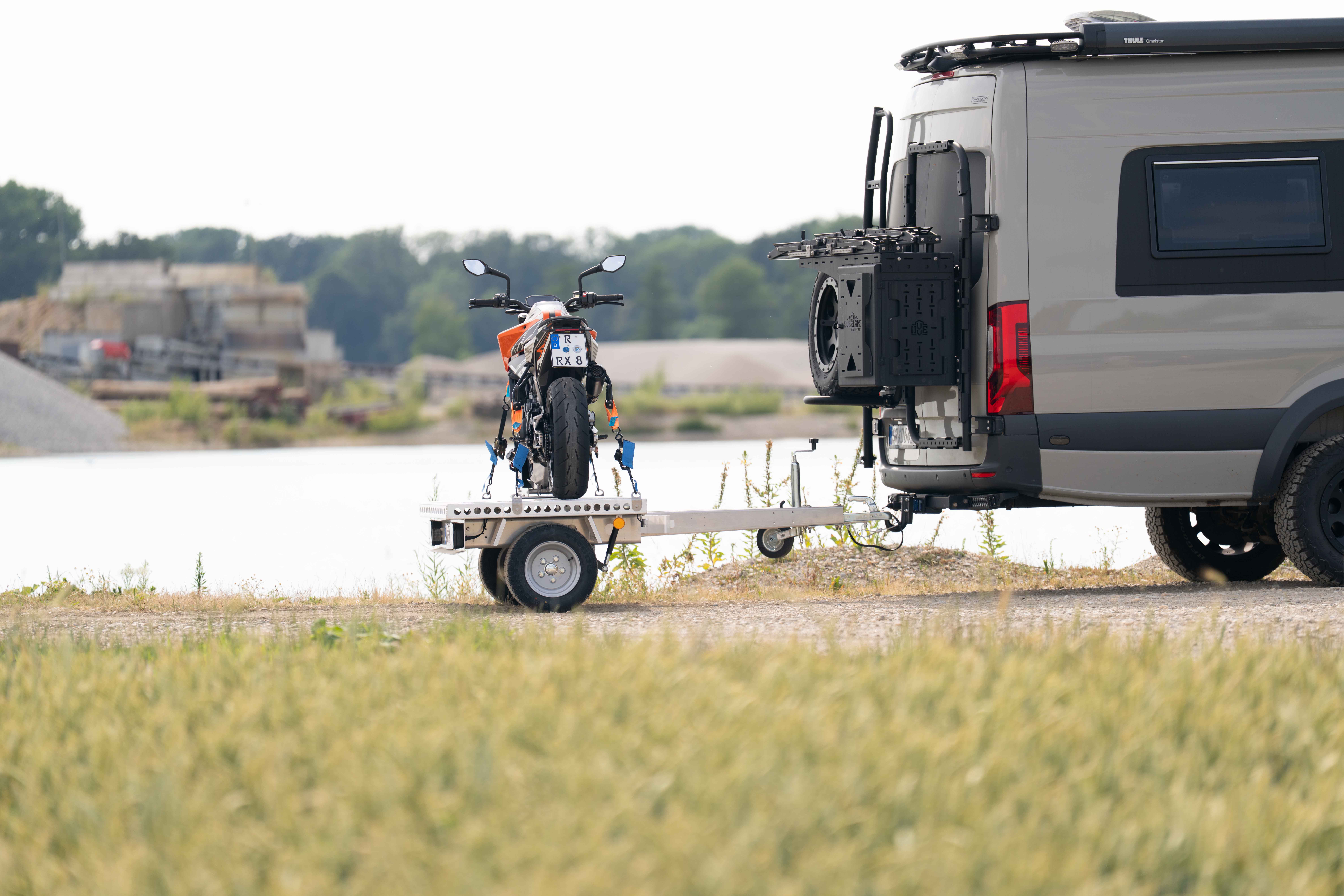 Kleiner Aluminium Plattformanhänger mit einer KTM Duke beladen hinter einem grünen Mercedes Sprinter auf einer grünen Wiese