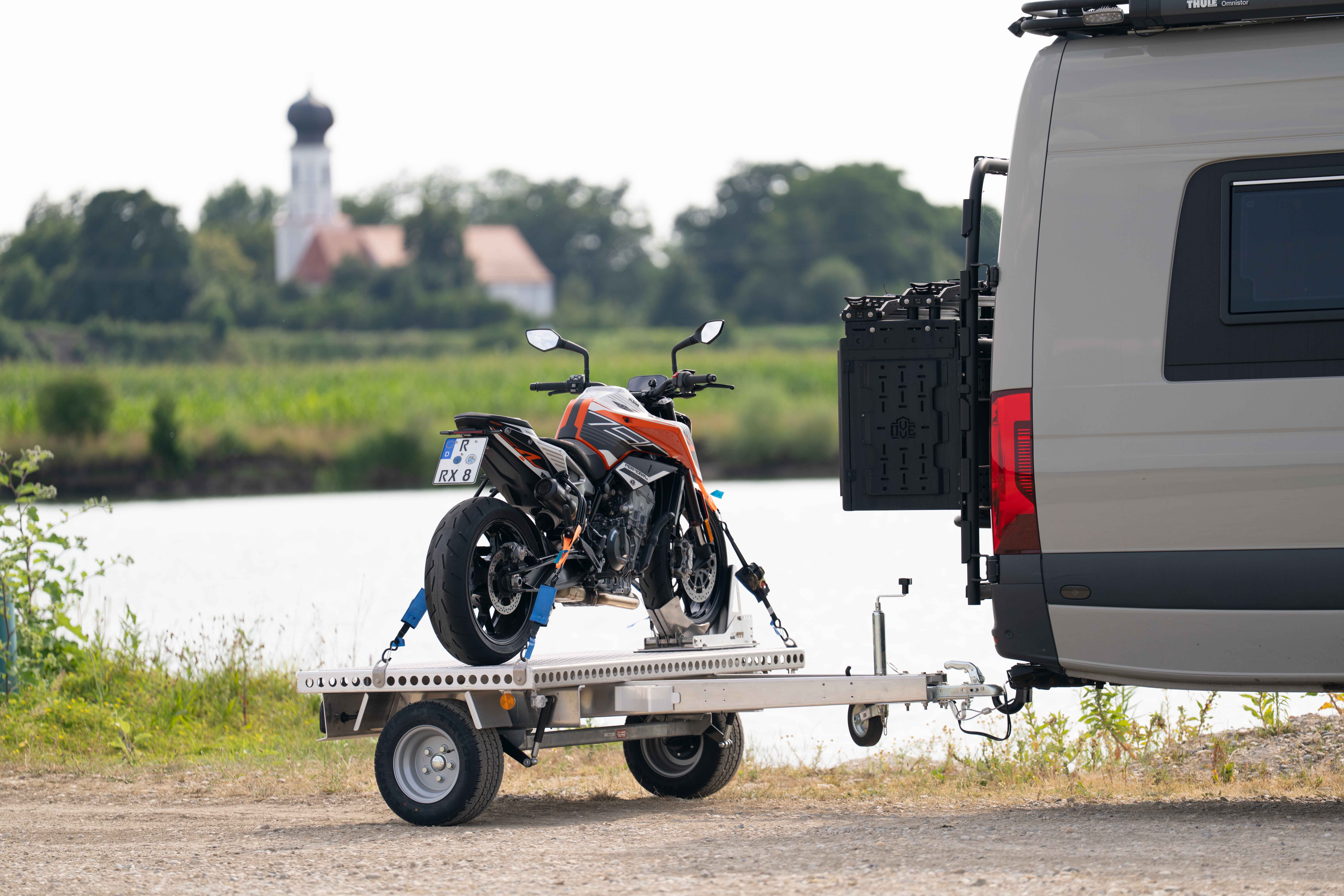 Kleiner Aluminium Plattformanhänger mit einer KTM Duke beladen hinter einem grünen Mercedes Sprinter auf einer grünen Wiese