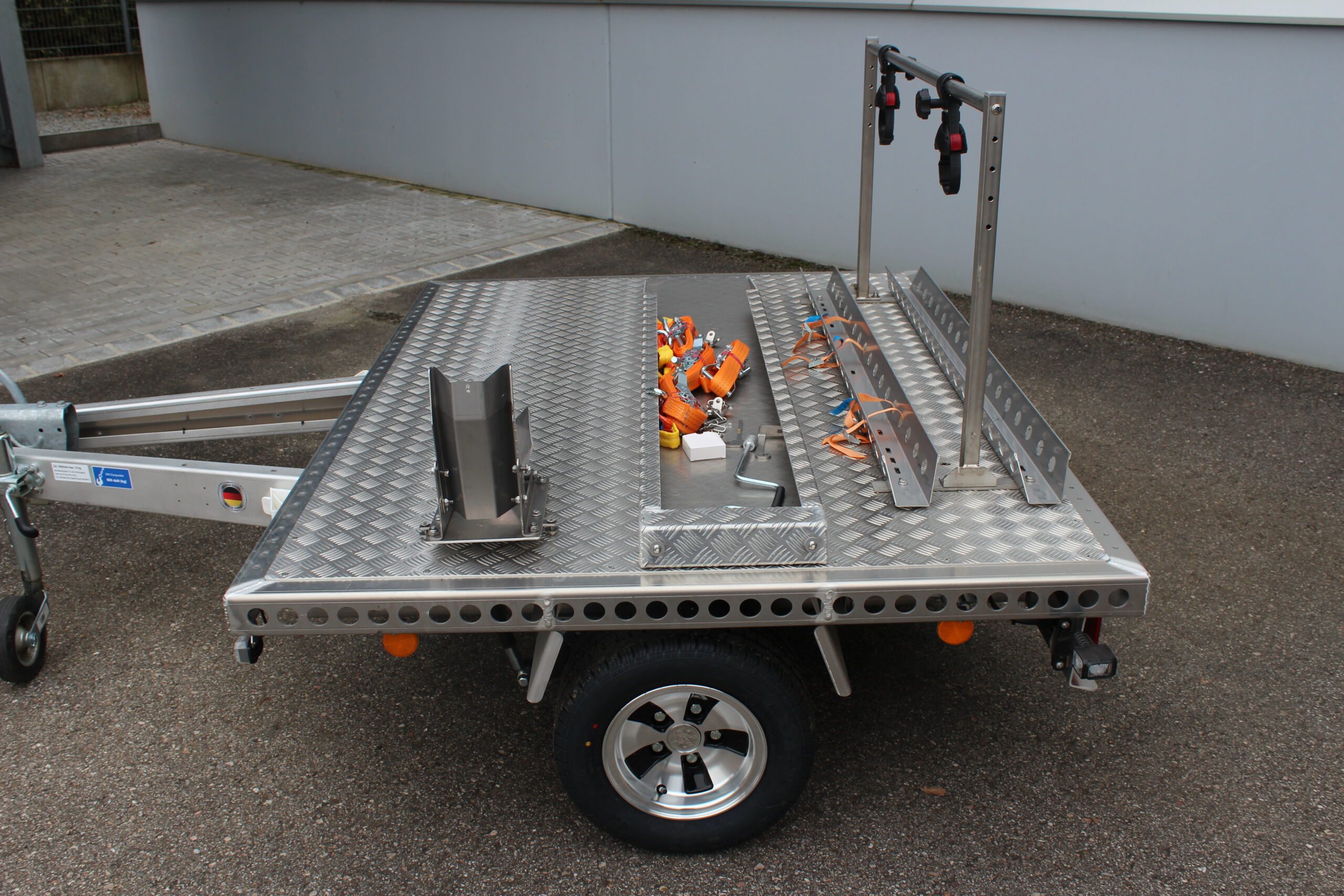 Aluminium Plattformanhänger mit Fahrradträger, Auffahrrampe und Radwippe vor einer grauen Wand