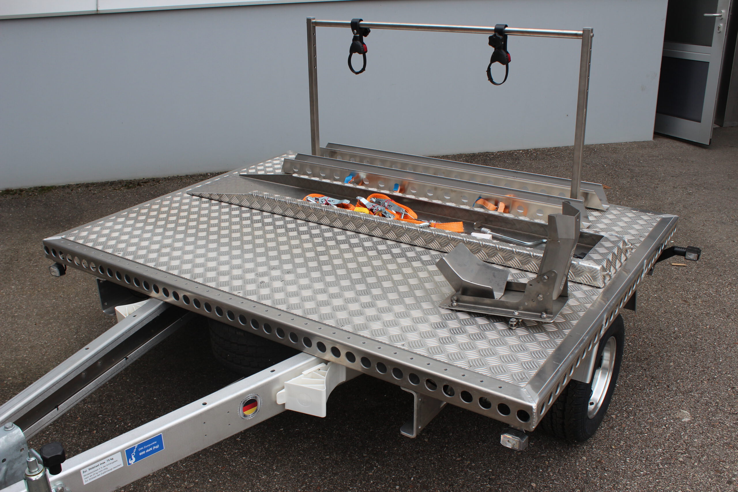 Aluminium Plattformanhänger mit Fahrradträger, Auffahrrampe und Radwippe vor einer grauen Wand