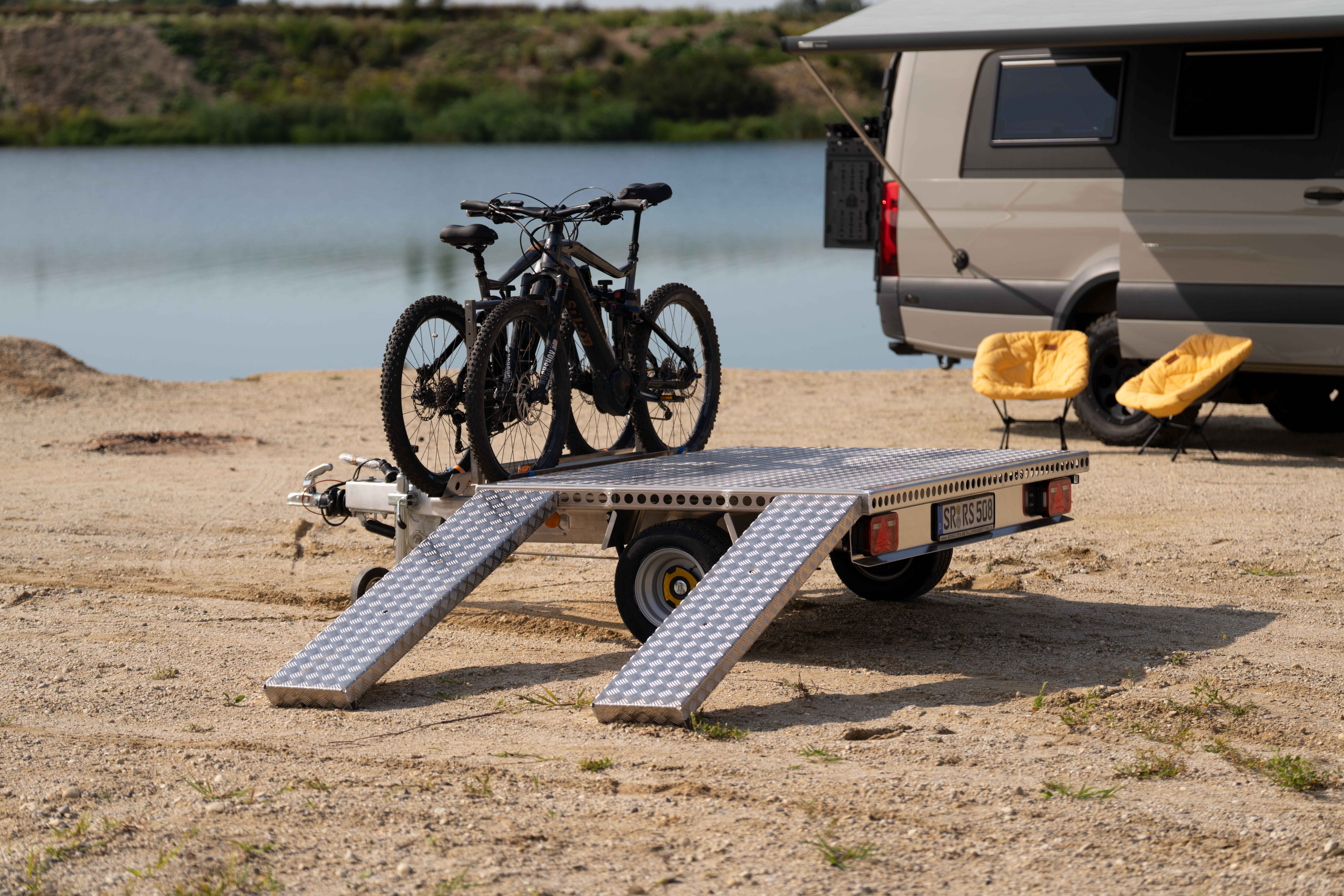 Aluminium Plattformanhänger mit zwei E-Bikes und zwei Auffahrrampen auf einer Sandfläche vor einem See