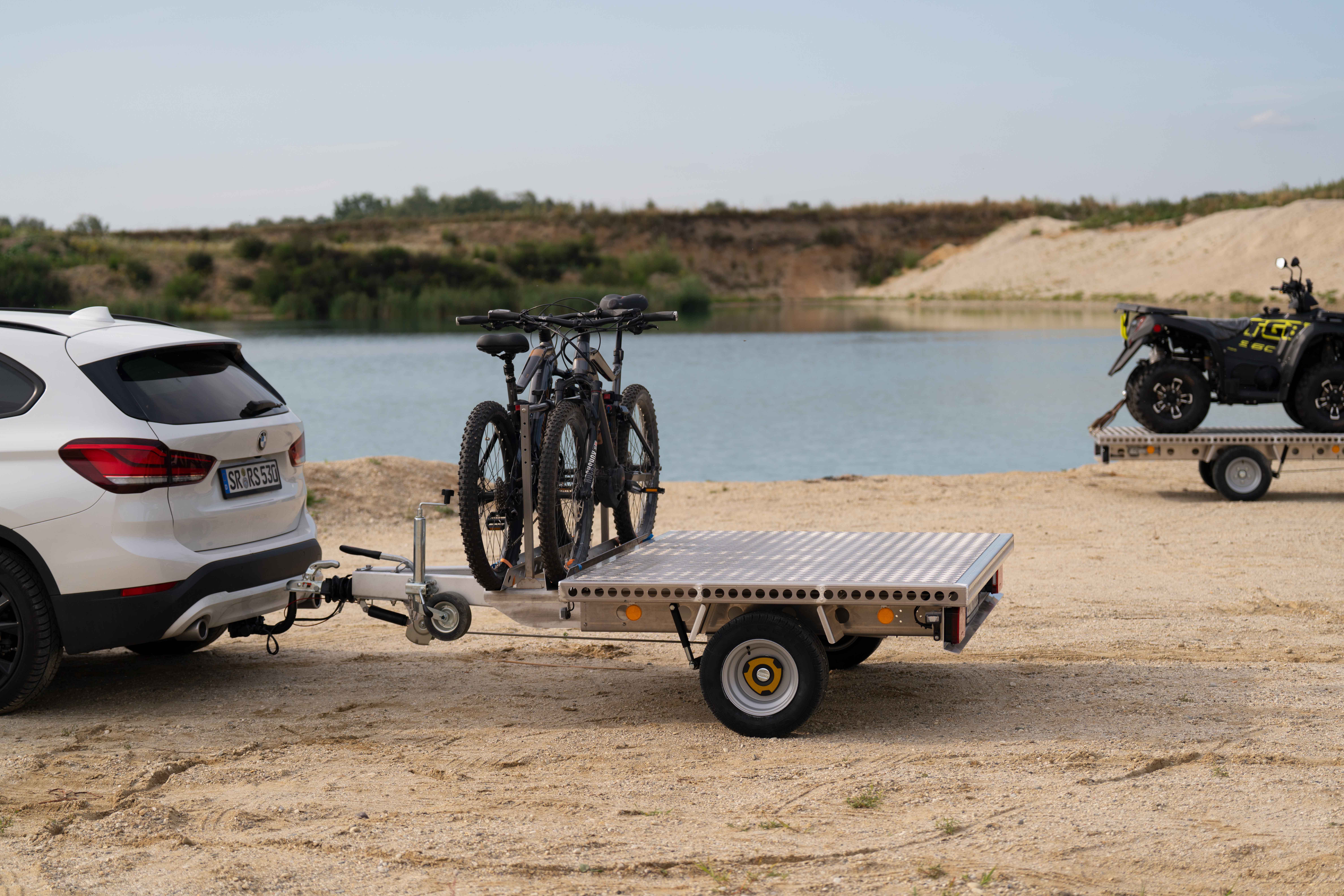Aluminium Plattformanhänger mit zwei E-Bikes auf einer Sandfläche vor einem See