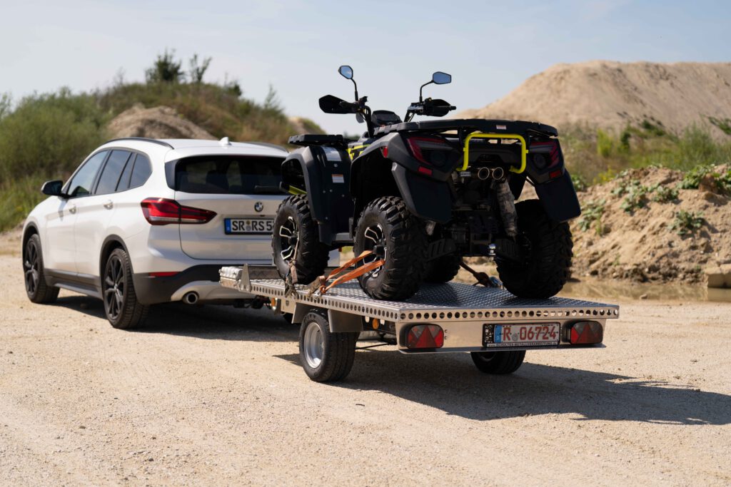 Aluminium Plattformanhänger mit einem Quad beladen hinter einem weißen Auto in einer Kiesgrube