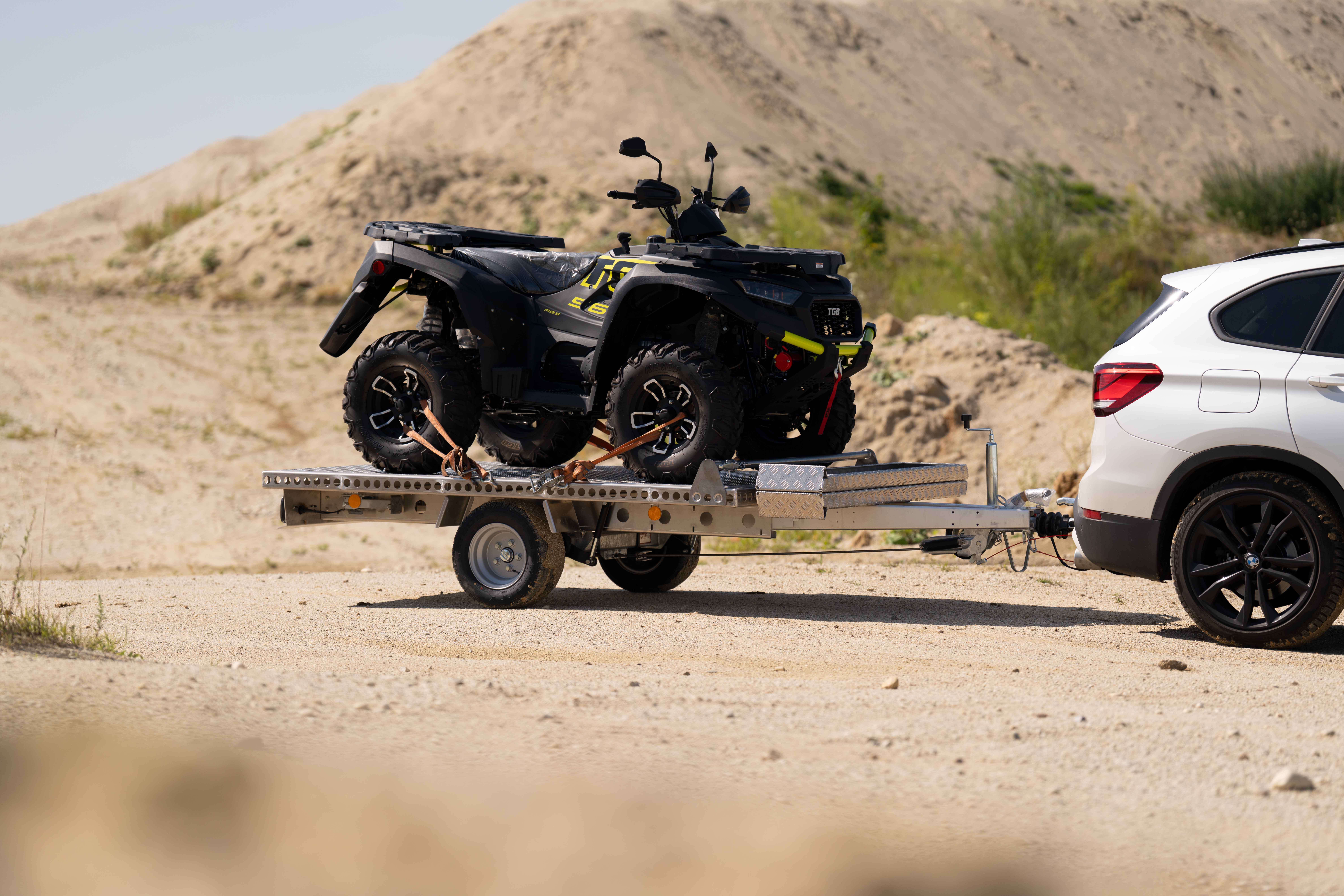 Aluminium Plattformanhänger mit einem Quad beladen hinter einem weißen Auto in einer Kiesgrube