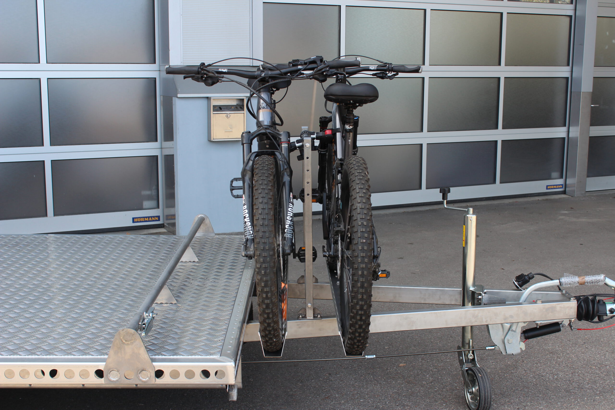 Fahrradträger für 2 Fahrräder aus Edelstahl an einem Anhänger befestigt mit 2 E-Bikes beladen vor einer Halle