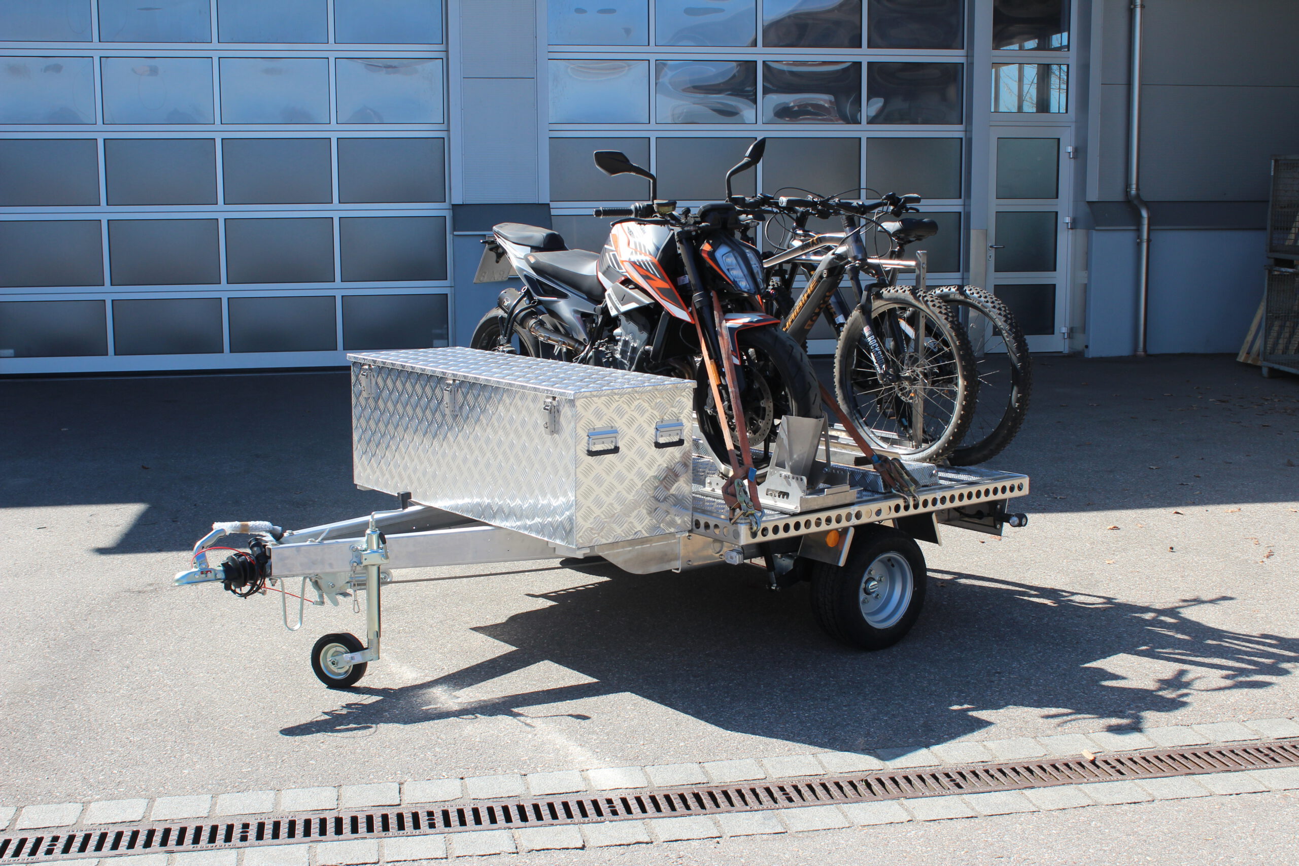UA150 Quer Anhänger mit einer KTM Duke und zwei Fahrrädern beladen. Zusätzlich ist eine Aluminium Staubox vorne quer verbaut. Vor einer Halle