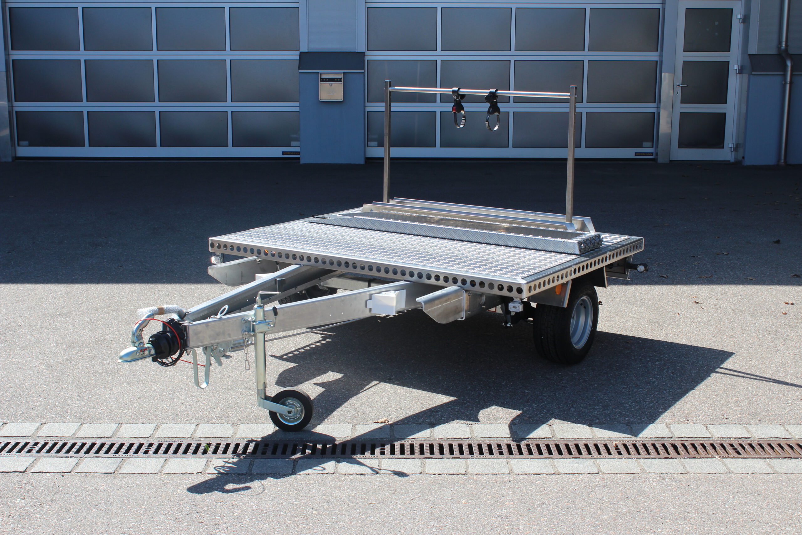 UA150 Quer Anhänger auf Aluminium mit einem Fahrradträger und einer Auffahrrampe auf der Plattform Vor einer Halle