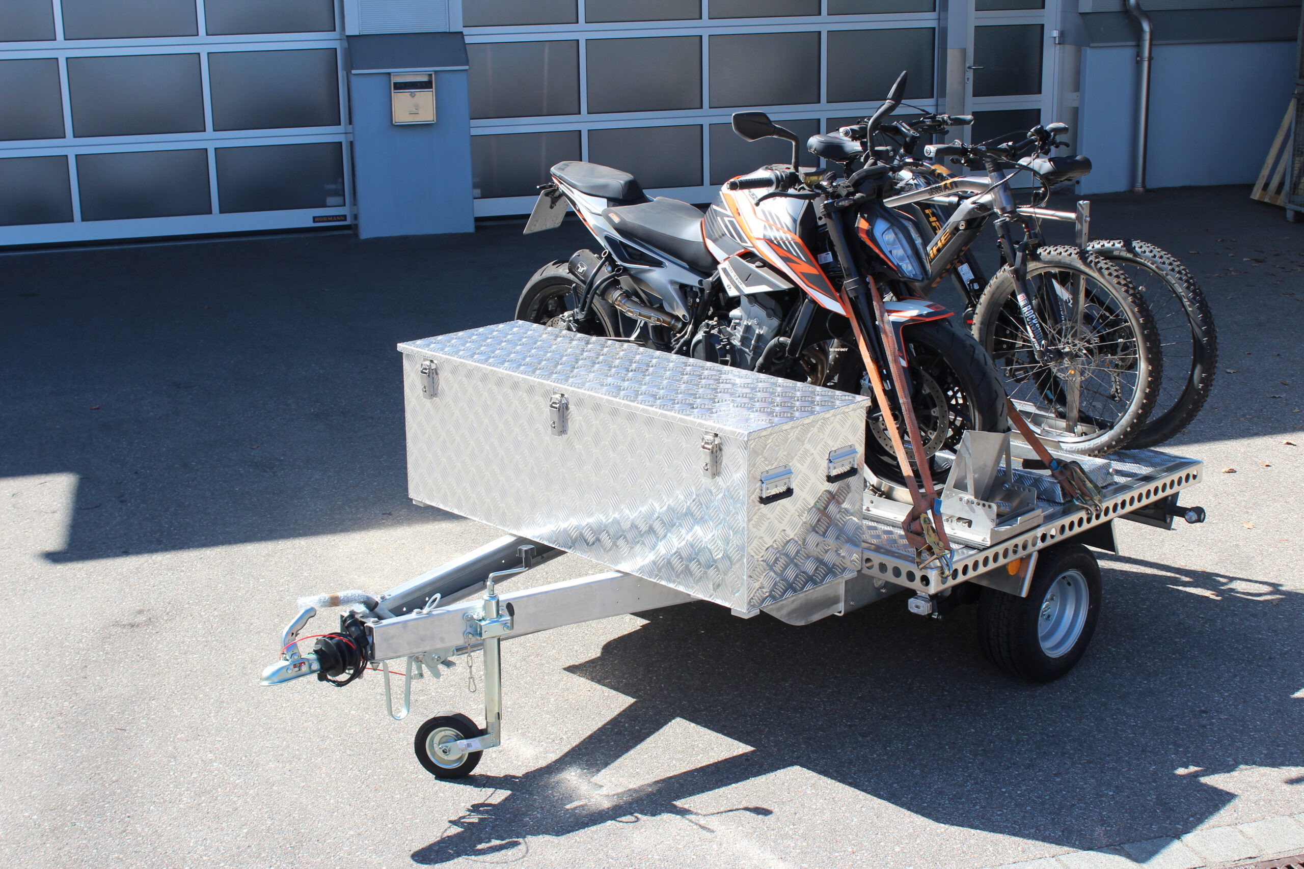 UA150 Quer Anhänger mit einer KTM Duke und zwei Fahrrädern beladen. Zusätzlich ist eine Aluminium Staubox vorne quer verbaut. Vor einer Halle