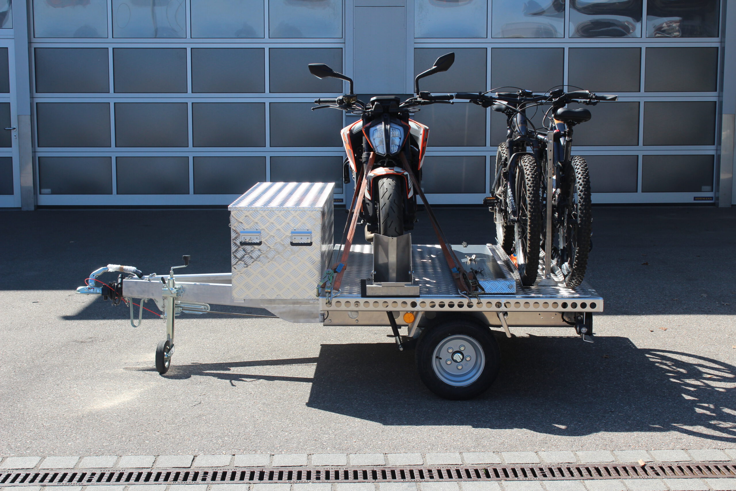 UA150 Quer Anhänger mit einer KTM Duke und zwei Fahrrädern beladen. Zusätzlich ist eine Aluminium Staubox vorne quer verbaut. Vor einer Halle