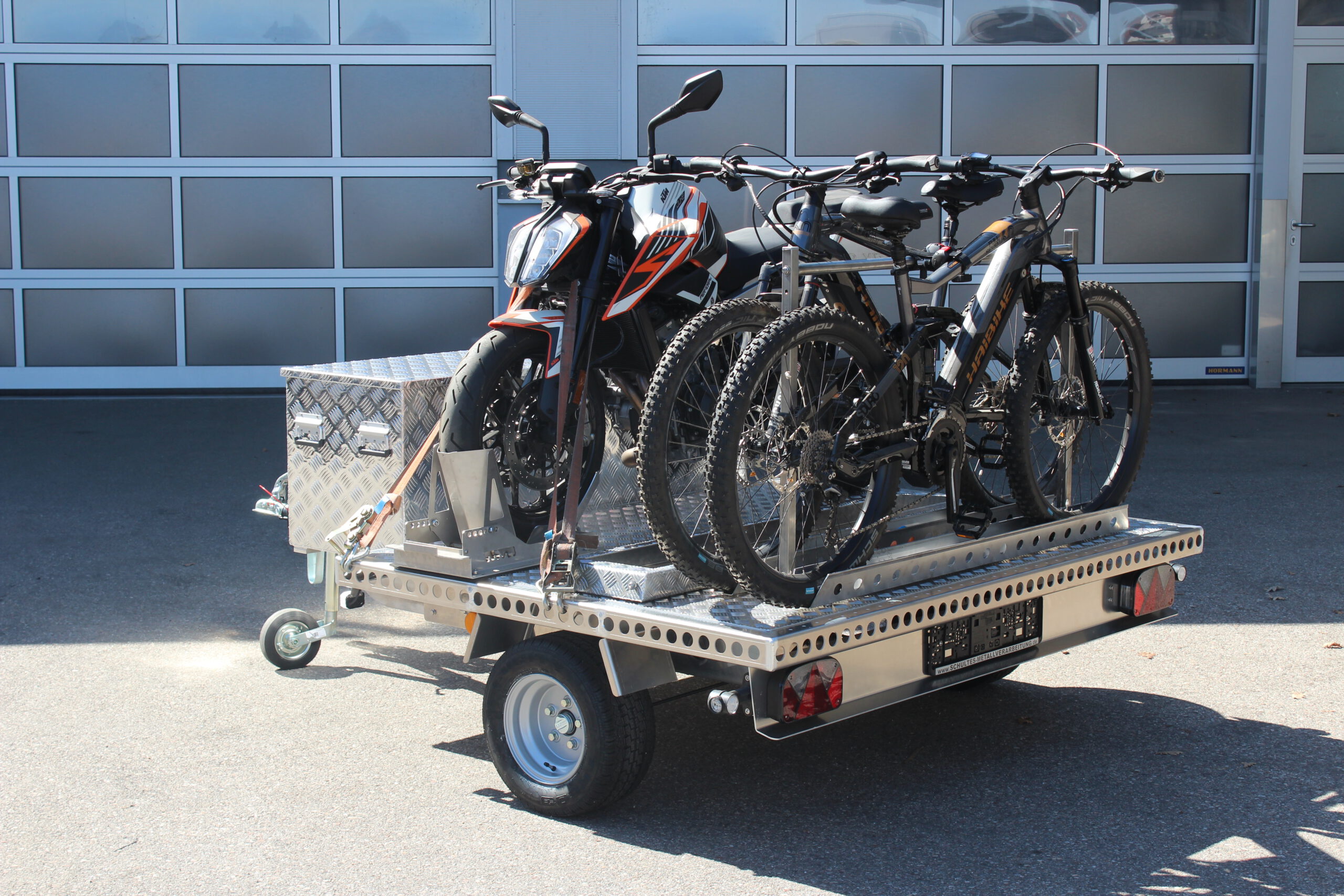 UA150 Quer Anhänger mit einer KTM Duke und zwei Fahrrädern beladen. Zusätzlich ist eine Aluminium Staubox vorne quer verbaut. Vor einer Halle