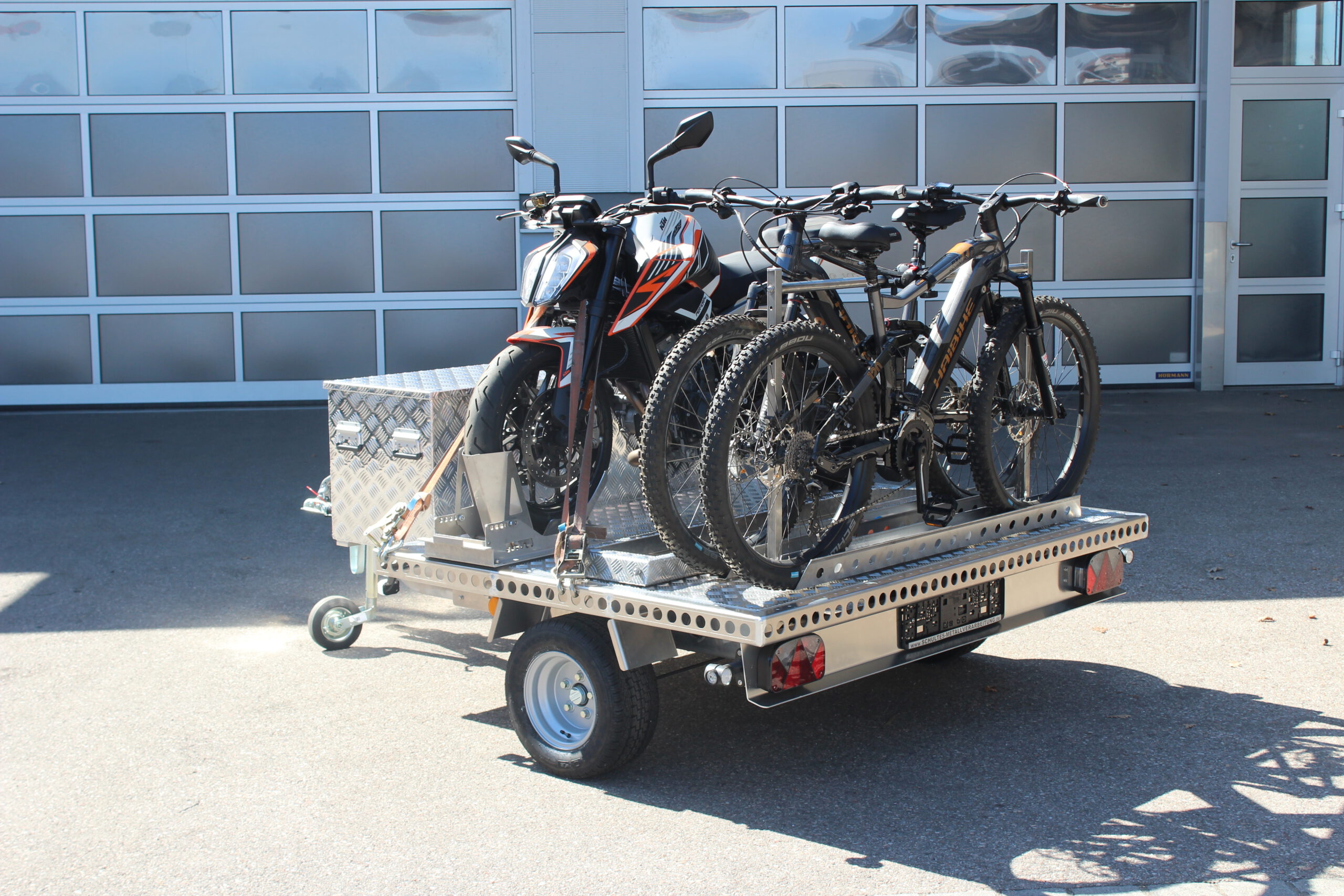 UA150 Quer Anhänger mit einer KTM Duke und zwei Fahrrädern beladen. Zusätzlich ist eine Aluminium Staubox vorne quer verbaut. Vor einer Halle