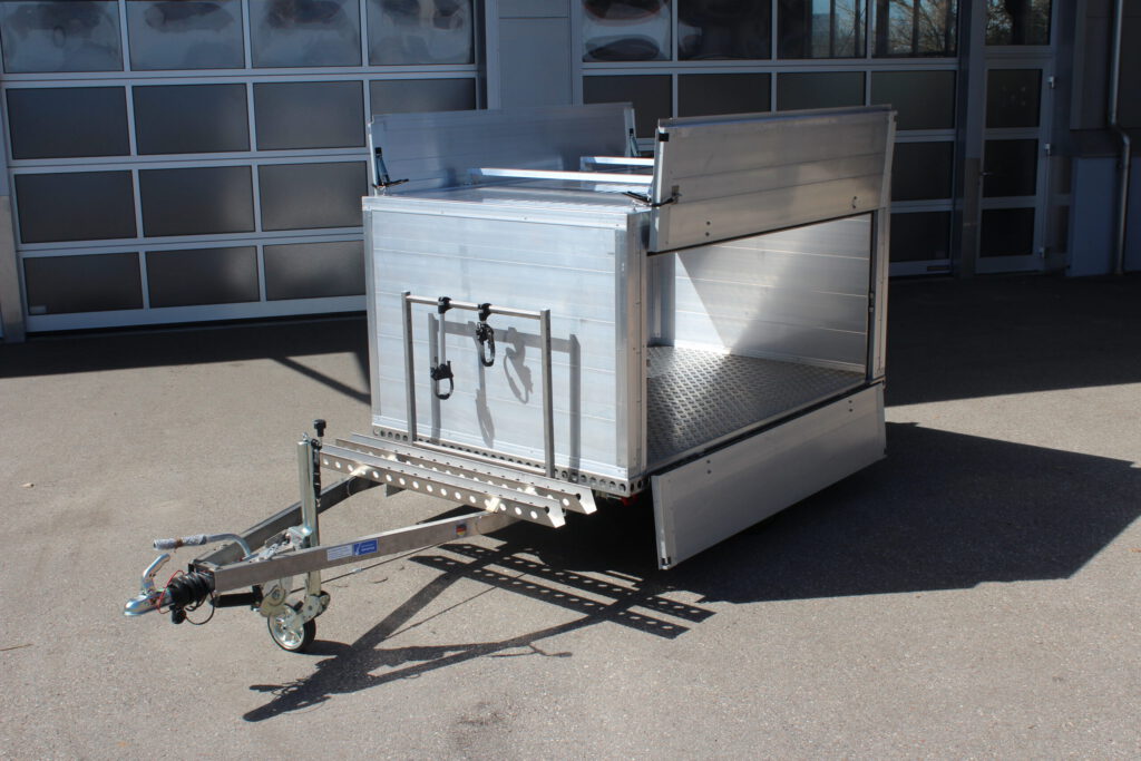 Aluminium Kofferanhänger mit einem Fahrradträger auf der Zugdeichsel. Aluminiumfelgen in Schwarz und ein Dachträger für ein Dachzelt vor einer Halle. Seitlich mit Klappen zum öffnen für das Be- und Entladen des Koffers