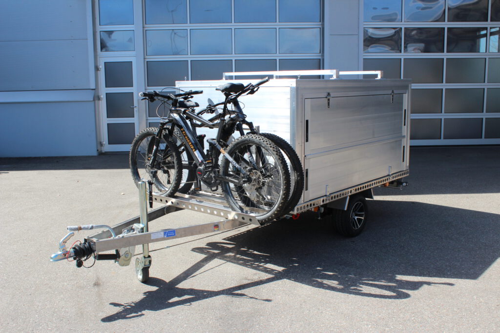 Aluminium Kofferanhänger mit einem Fahrradträger auf der Zugdeichsel. Aluminiumfelgen in Schwarz und ein Dachträger für ein Dachzelt vor einer Halle. Seitlich mit Klappen zum öffnen für das Be- und Entladen des Koffers
