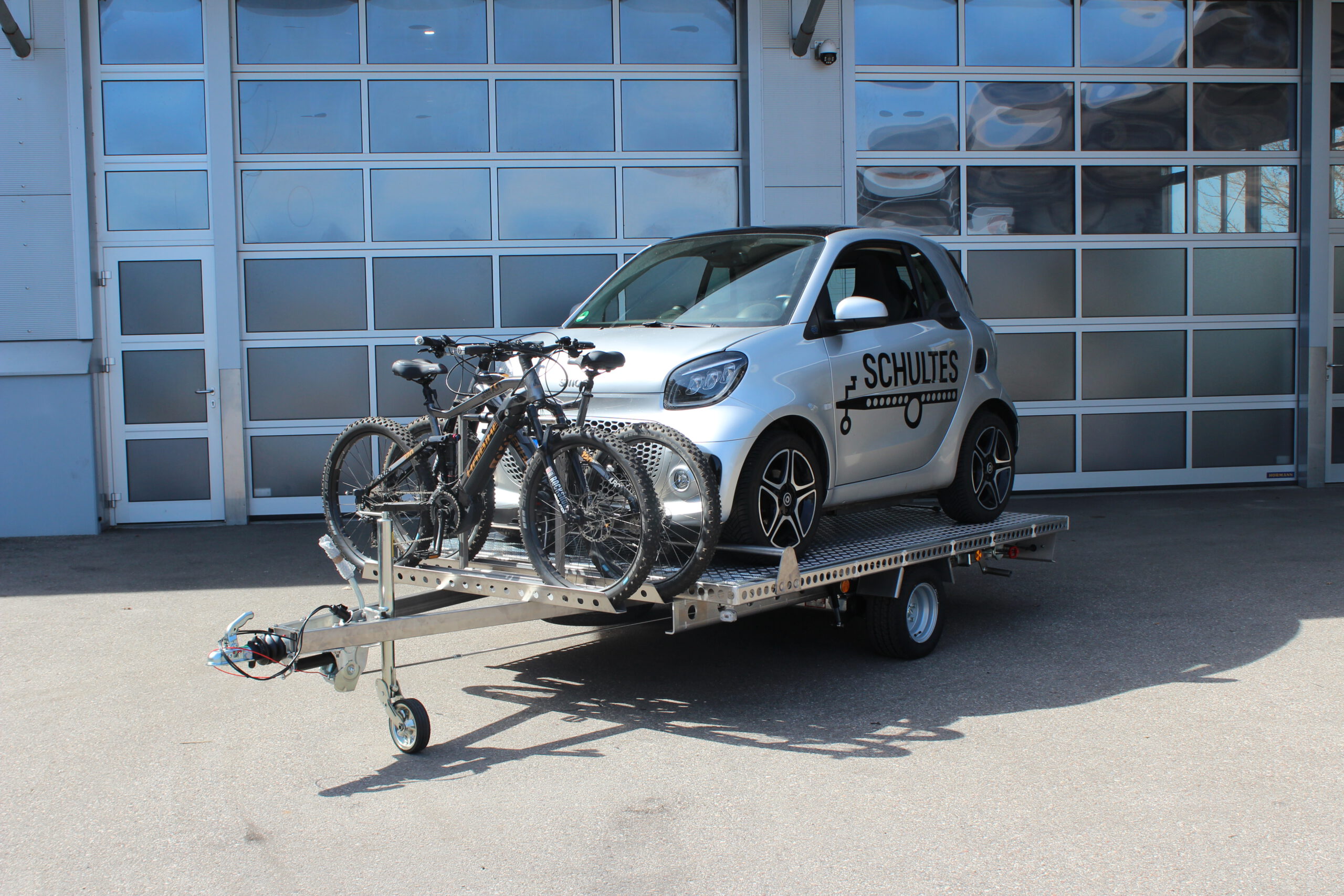 Aluminium Anhänger mit einem Smart beladen und einem Edelstahl Fahrradträger mit 2 E-Bikes auf der Zugdeichsel vor einer Halle