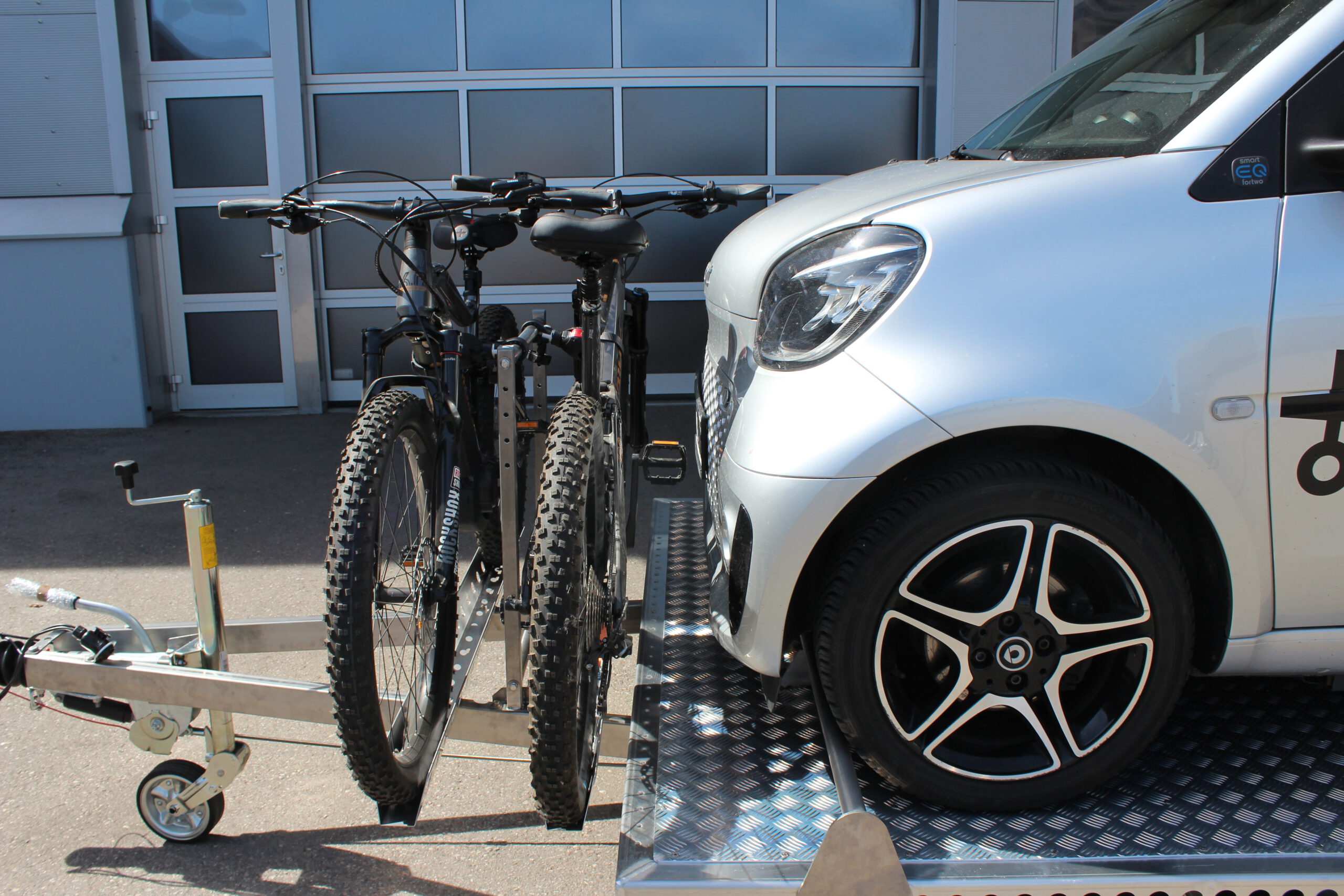 Aluminium Anhänger mit einem Smart beladen und einem Edelstahl Fahrradträger mit 2 E-Bikes auf der Zugdeichsel vor einer Halle
