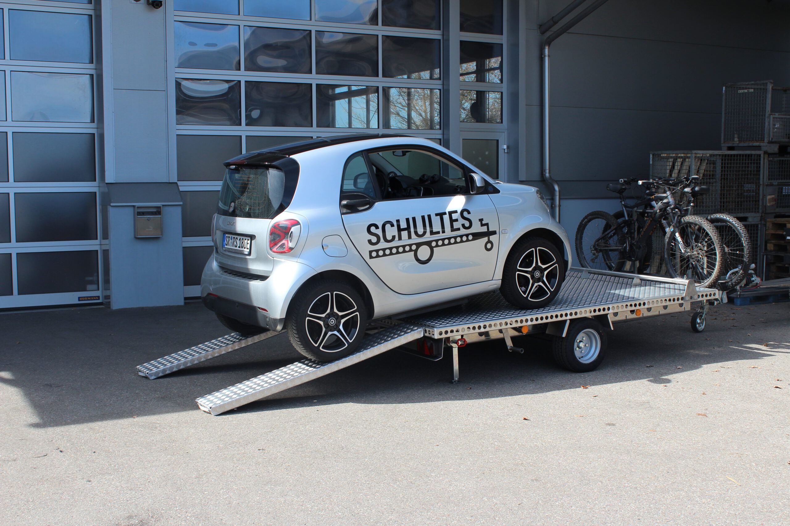 Aluminium Anhänger mit einem Smart beladen und einem Edelstahl Fahrradträger mit 2 E-Bikes auf der Zugdeichsel vor einer Halle
