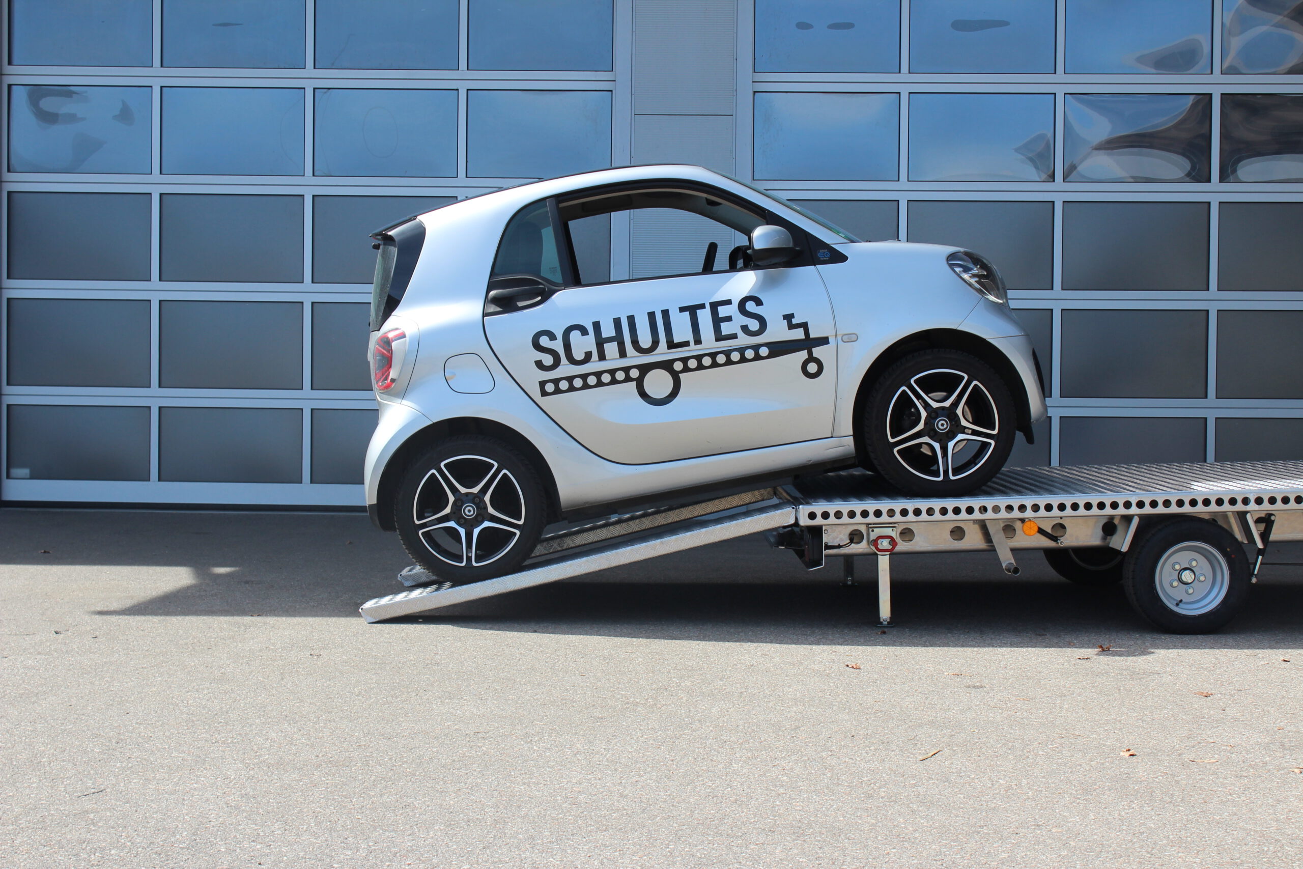 Aluminium Anhänger mit einem Smart beladen und einem Edelstahl Fahrradträger mit 2 E-Bikes auf der Zugdeichsel vor einer Halle