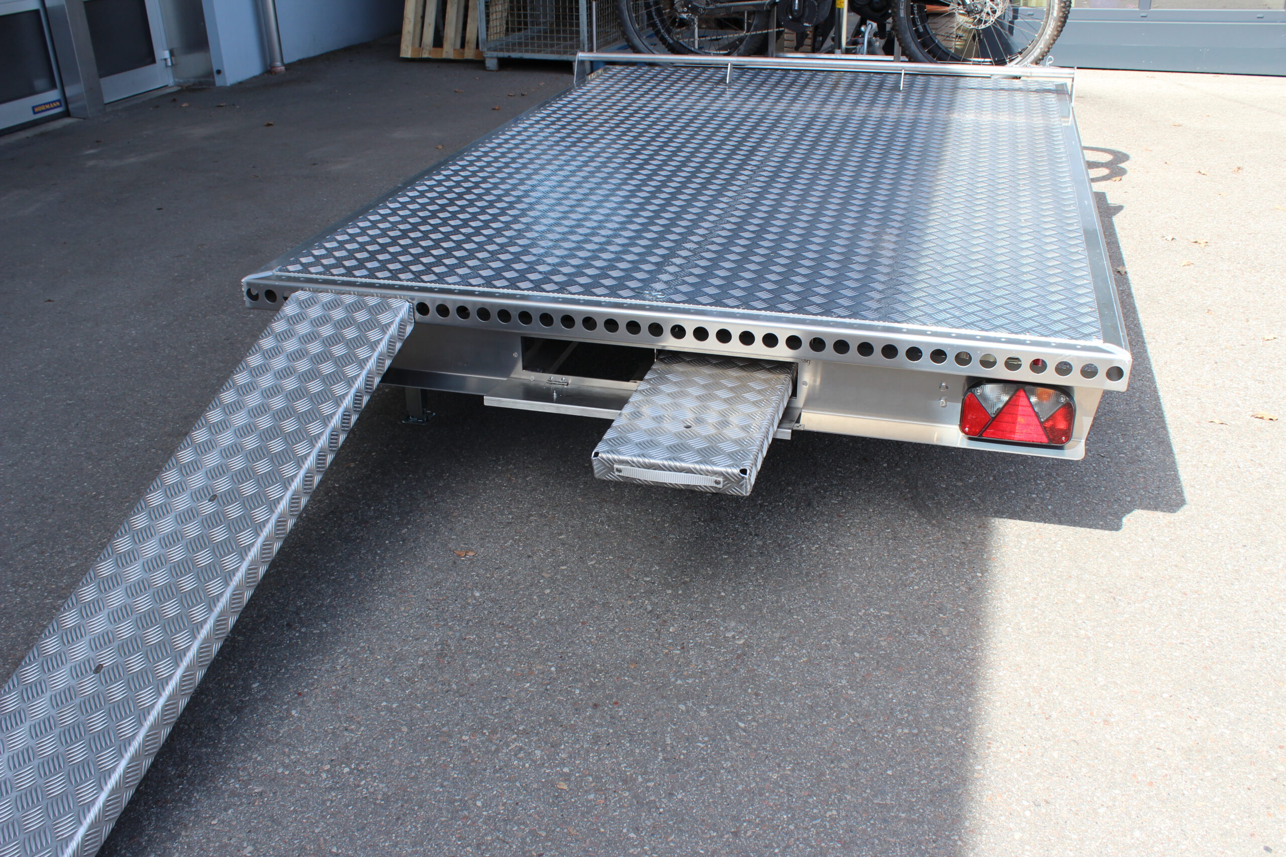Aluminium Anhänger mit einem Edelstahl Fahrradträger mit 2 E-Bikes auf der Zugdeichsel vor einer Halle