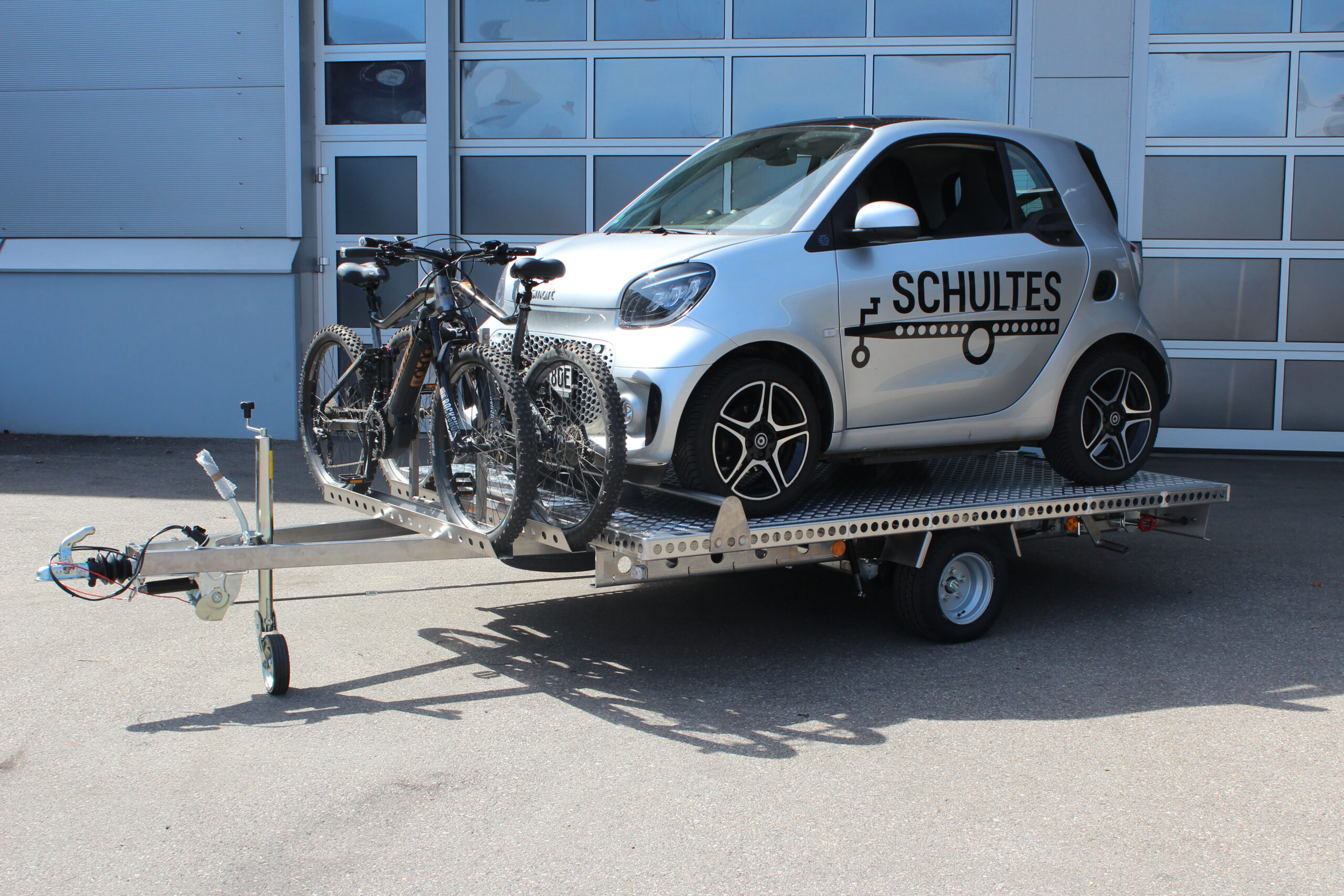 Aluminium Anhänger mit einem Smart beladen und einem Edelstahl Fahrradträger mit 2 E-Bikes auf der Zugdeichsel vor einer Halle