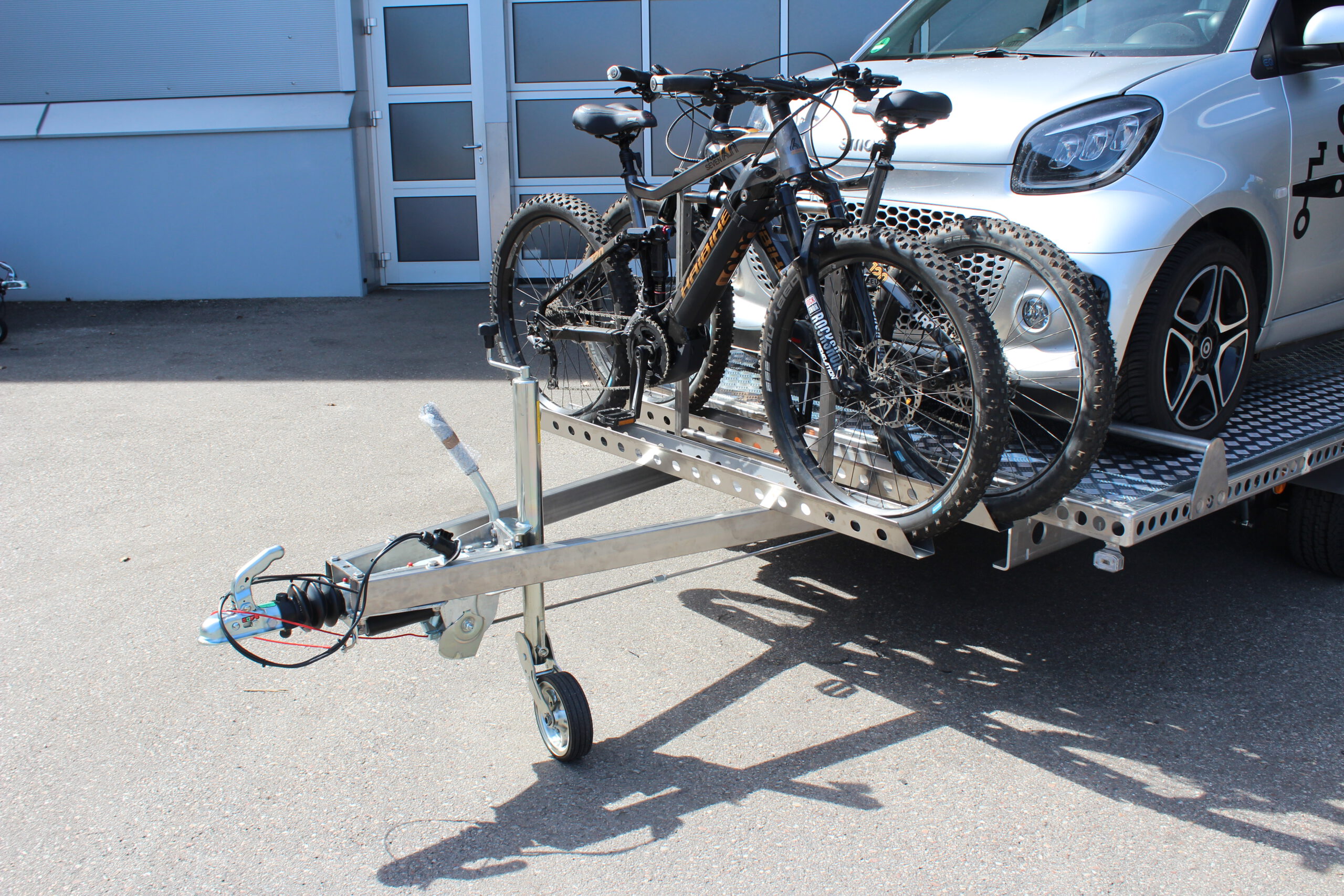 Aluminium Anhänger mit einem Smart beladen und einem Edelstahl Fahrradträger mit 2 E-Bikes auf der Zugdeichsel vor einer Halle