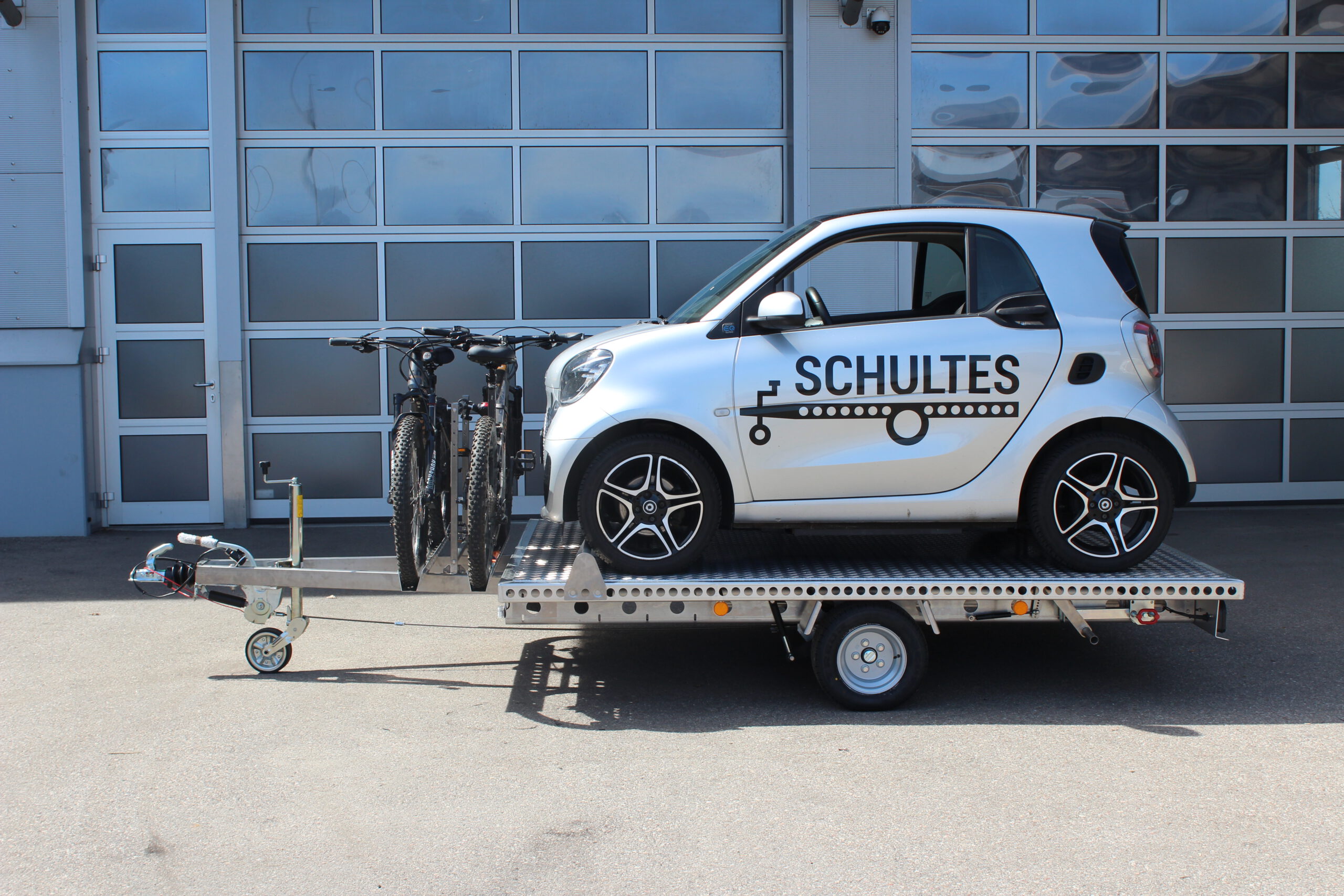 Aluminium Anhänger mit einem Smart beladen und einem Edelstahl Fahrradträger mit 2 E-Bikes auf der Zugdeichsel vor einer Halle