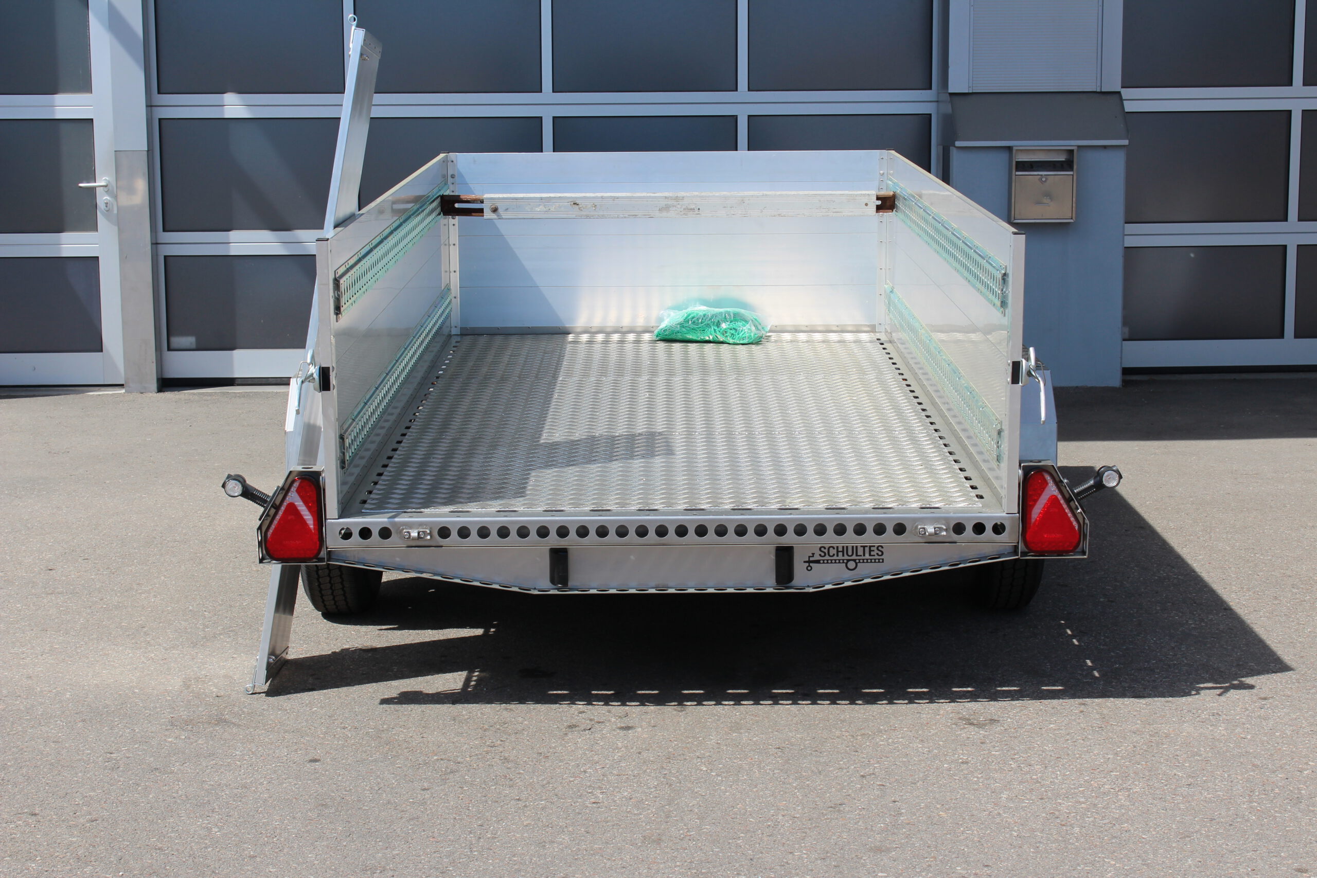 Edelstahl Tieflader Anhänger mit Aluminium Bordwänden für den gewerblichen Einsatz. Zweiachser aus Edelstahl mit Heckklappe vor einer Halle auf einem Hof