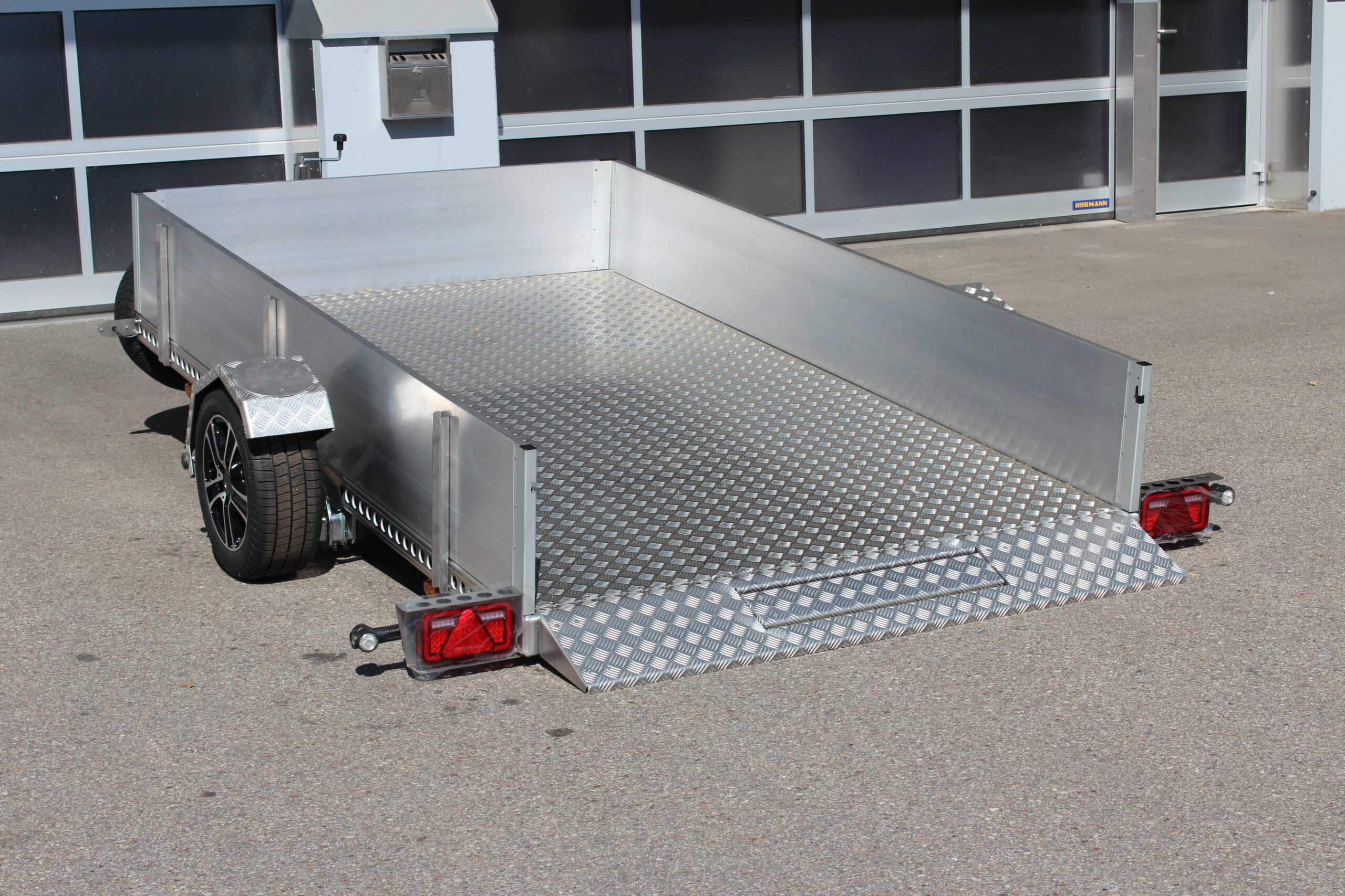 AS300 Absenkanhänger aus Aluminium mit Bordwänden aus Aluminium 40cm hoch mit manueller Handpumpe, abgesenkt auf einem Hof vor einem Hallentor, Ansicht von hinten