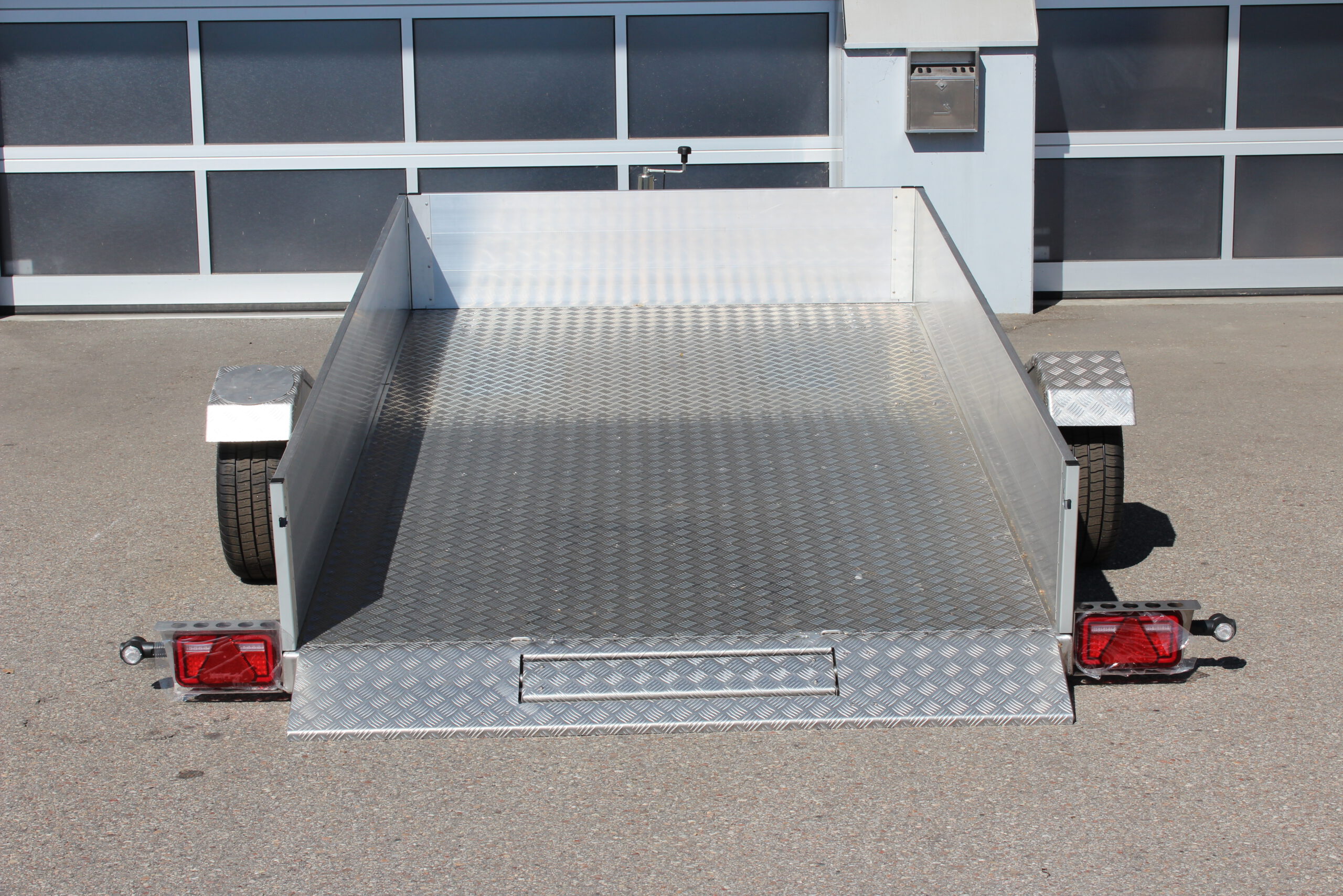 AS300 Absenkanhänger aus Aluminium mit Bordwänden aus Aluminium 40cm hoch mit manueller Handpumpe, abgesenkt auf einem Hof vor einem Hallentor, Ansicht von hinten