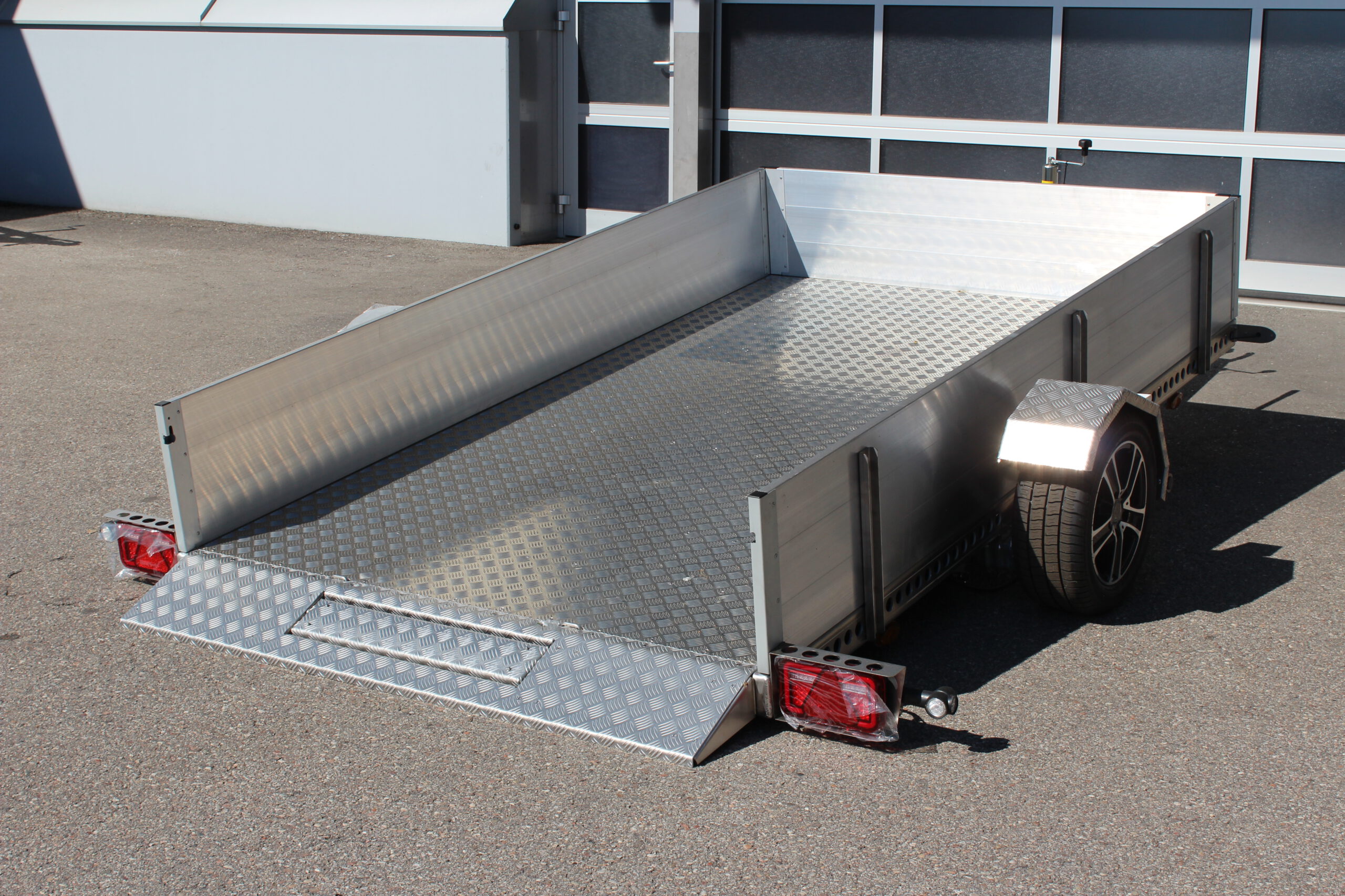 AS300 Absenkanhänger aus Aluminium mit Bordwänden aus Aluminium 40cm hoch mit manueller Handpumpe, abgesenkt auf einem Hof vor einem Hallentor, Ansicht von hinten