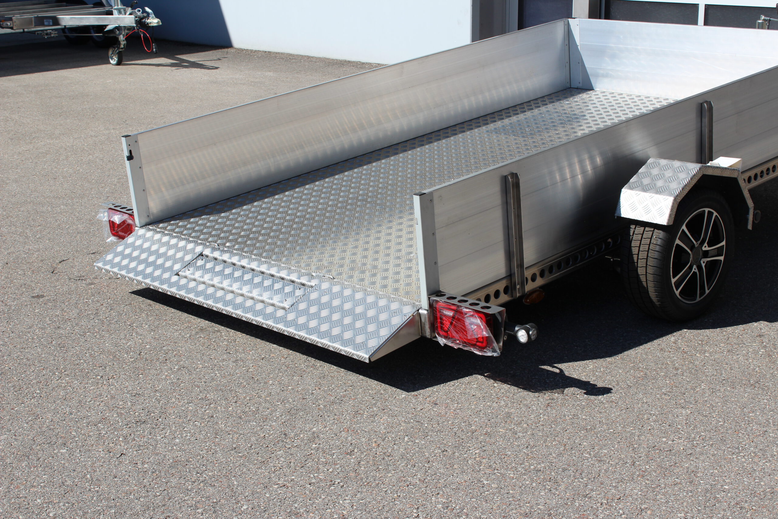 AS300 Absenkanhänger aus Aluminium mit Bordwänden aus Aluminium 40cm hoch mit manueller Handpumpe, abgesenkt auf einem Hof vor einem Hallentor, Ansicht von hinten