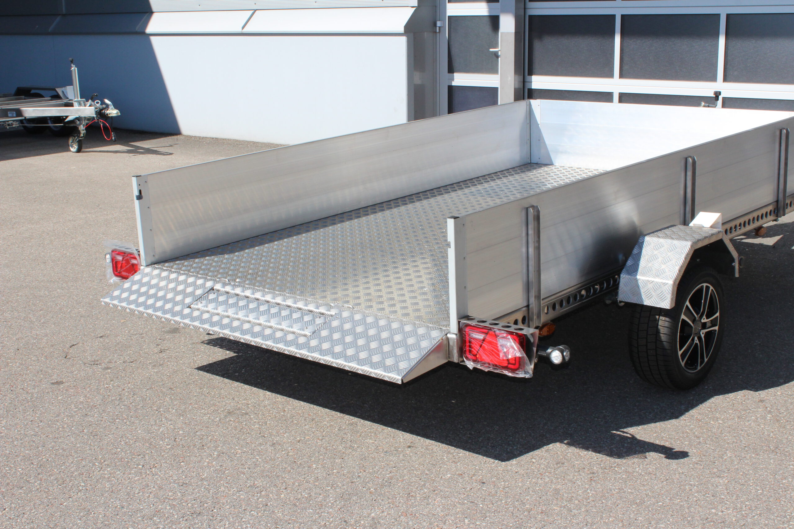 AS300 Absenkanhänger aus Aluminium mit Bordwänden aus Aluminium 40cm hoch mit manueller Handpumpe, auf einem Hof vor einem Hallentor, Ansicht von hinten