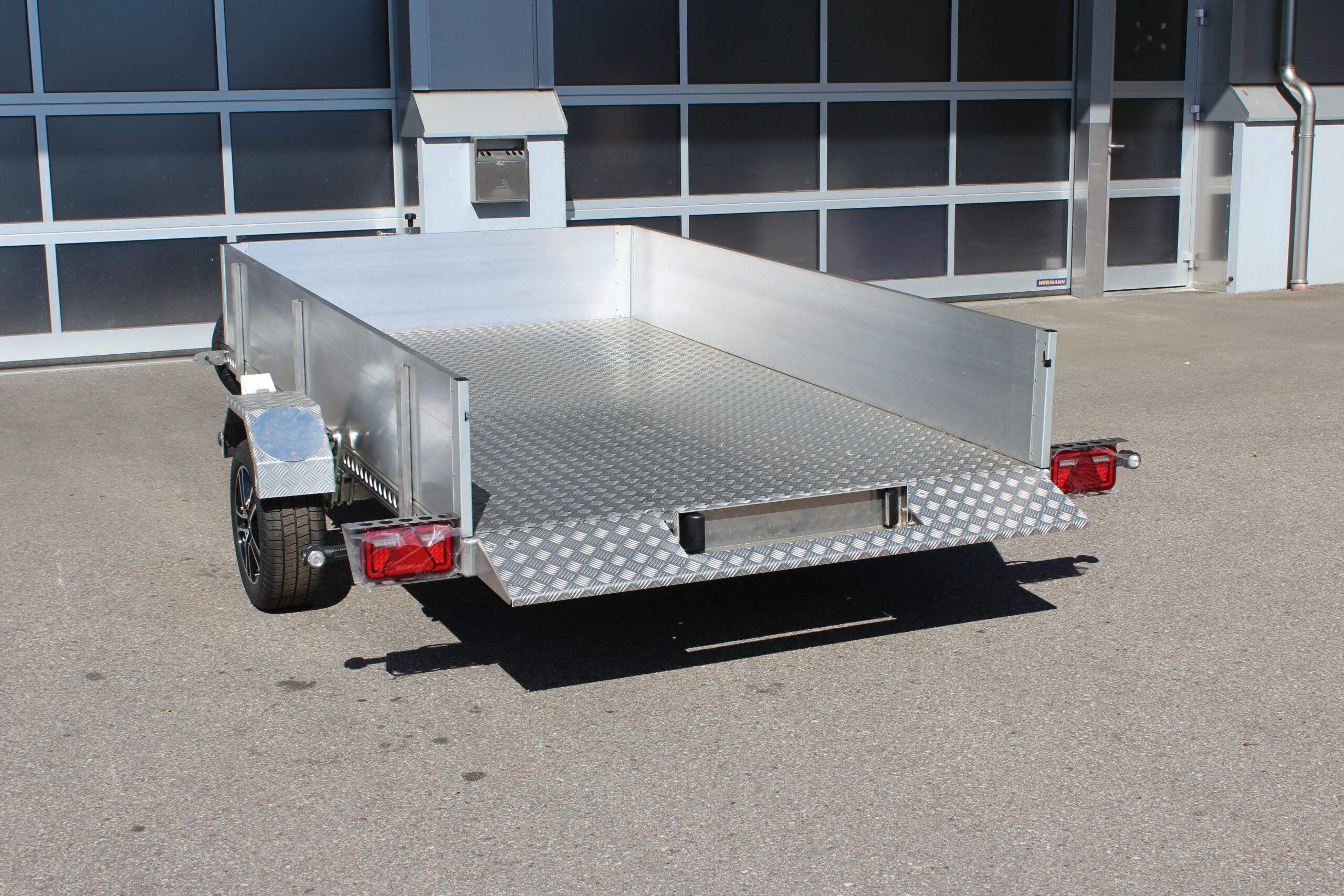 AS300 Absenkanhänger aus Aluminium mit Bordwänden aus Aluminium 40cm hoch mit manueller Handpumpe, auf einem Hof vor einem Hallentor, Ansicht von hinten