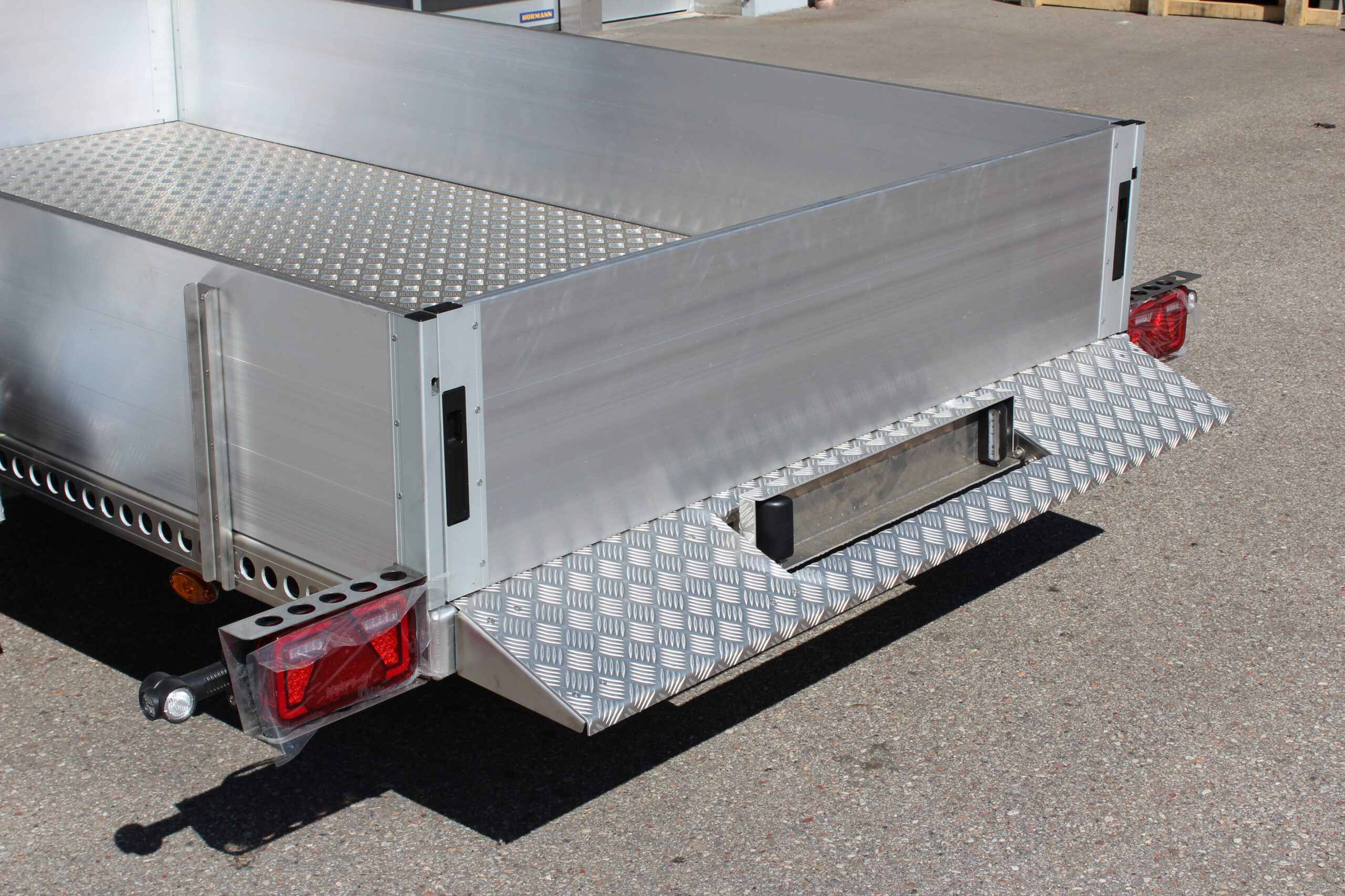 AS300 Absenkanhänger aus Aluminium mit Bordwänden aus Aluminium 40cm hoch mit manueller Handpumpe, auf einem Hof vor einem Hallentor, Ansicht von hinten