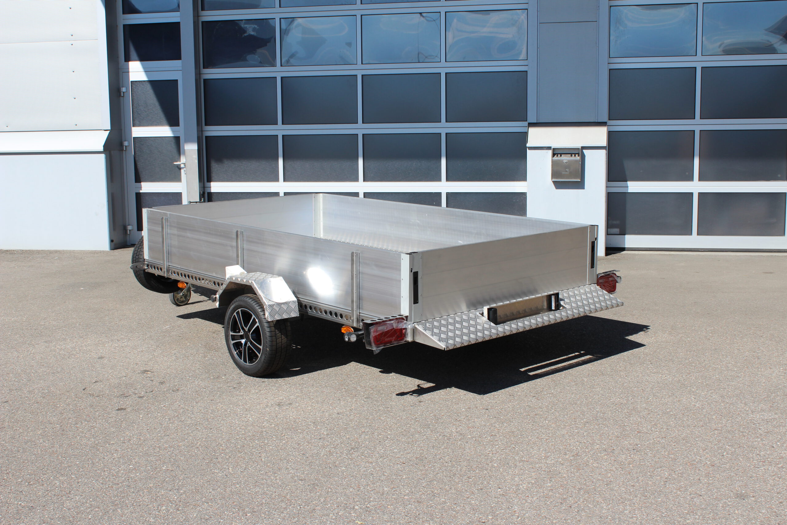 AS300 Absenkanhänger aus Aluminium mit Bordwänden aus Aluminium 40cm hoch mit manueller Handpumpe, auf einem Hof vor einem Hallentor, Ansicht von hinten