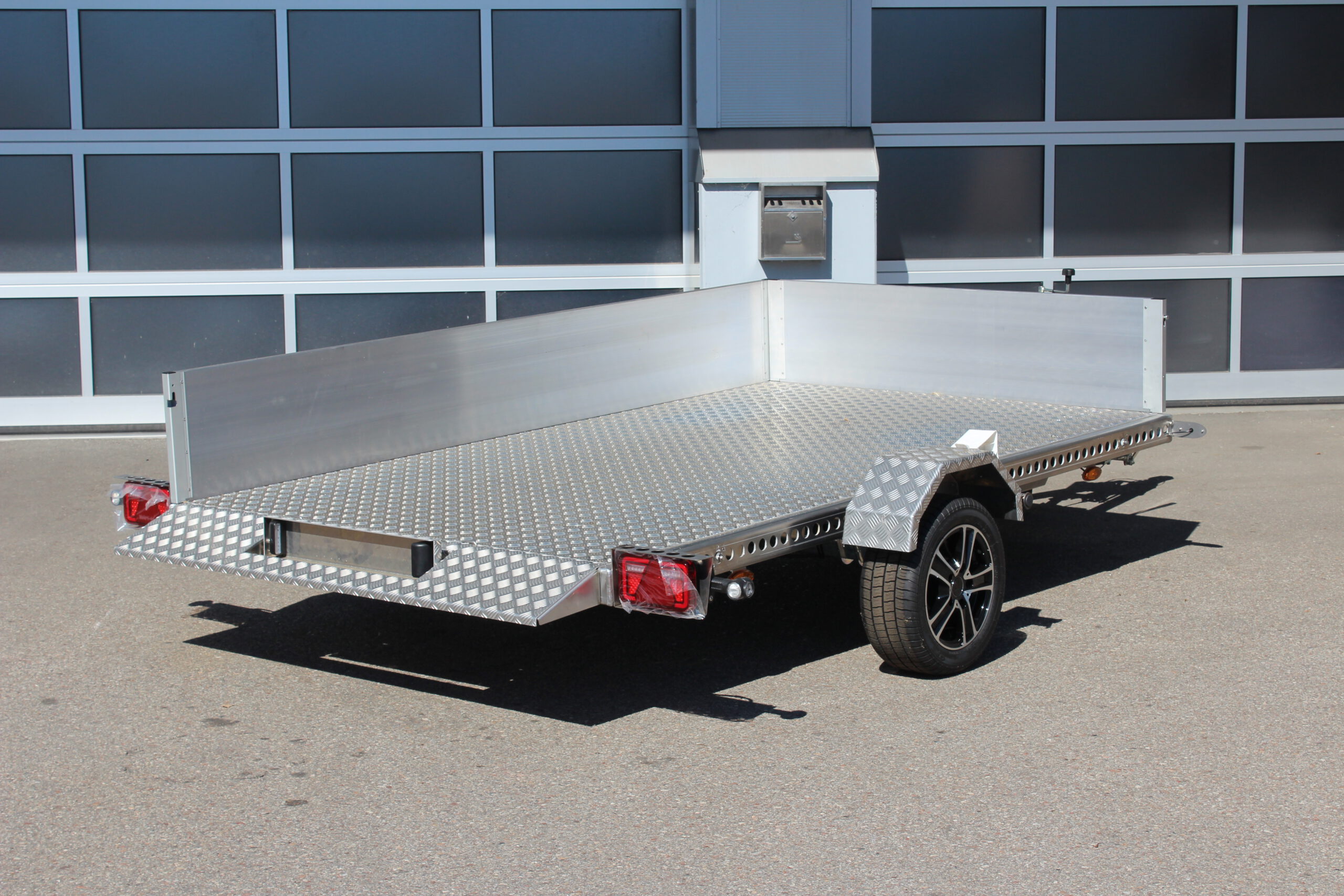 AS300 Absenkanhänger aus Aluminium mit Bordwänden aus Aluminium 40cm hoch mit manueller Handpumpe, auf einem Hof vor einem Hallentor, Ansicht von hinten