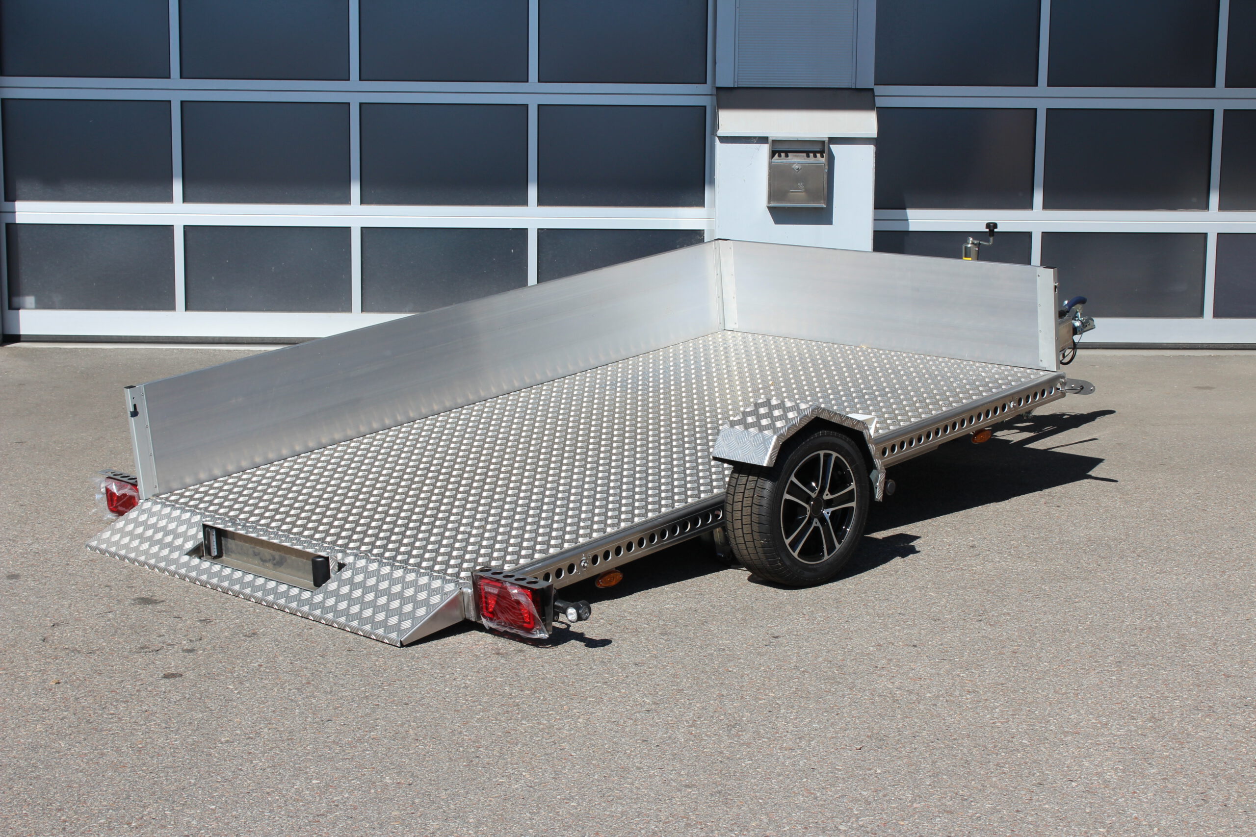 AS300 Absenkanhänger aus Aluminium mit Bordwänden aus Aluminium 40cm hoch mit manueller Handpumpe, auf einem Hof vor einem Hallentor, Ansicht von hinten