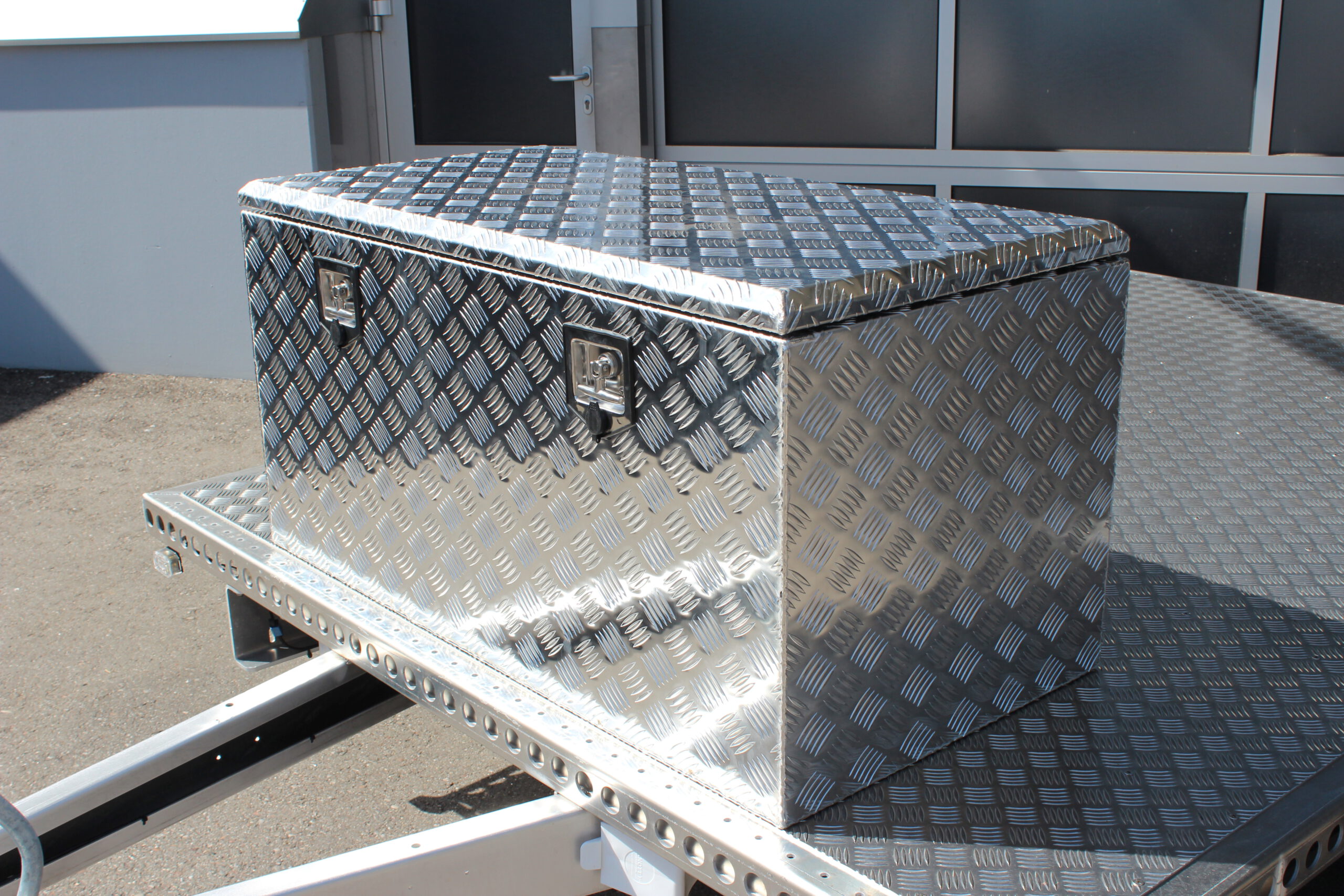 Große silberne Aluminium Riffelblech Staubox mit zwei Drehverschlüssen. Steht auf einem silbernen Aluminium Anhänger vor einem Tor