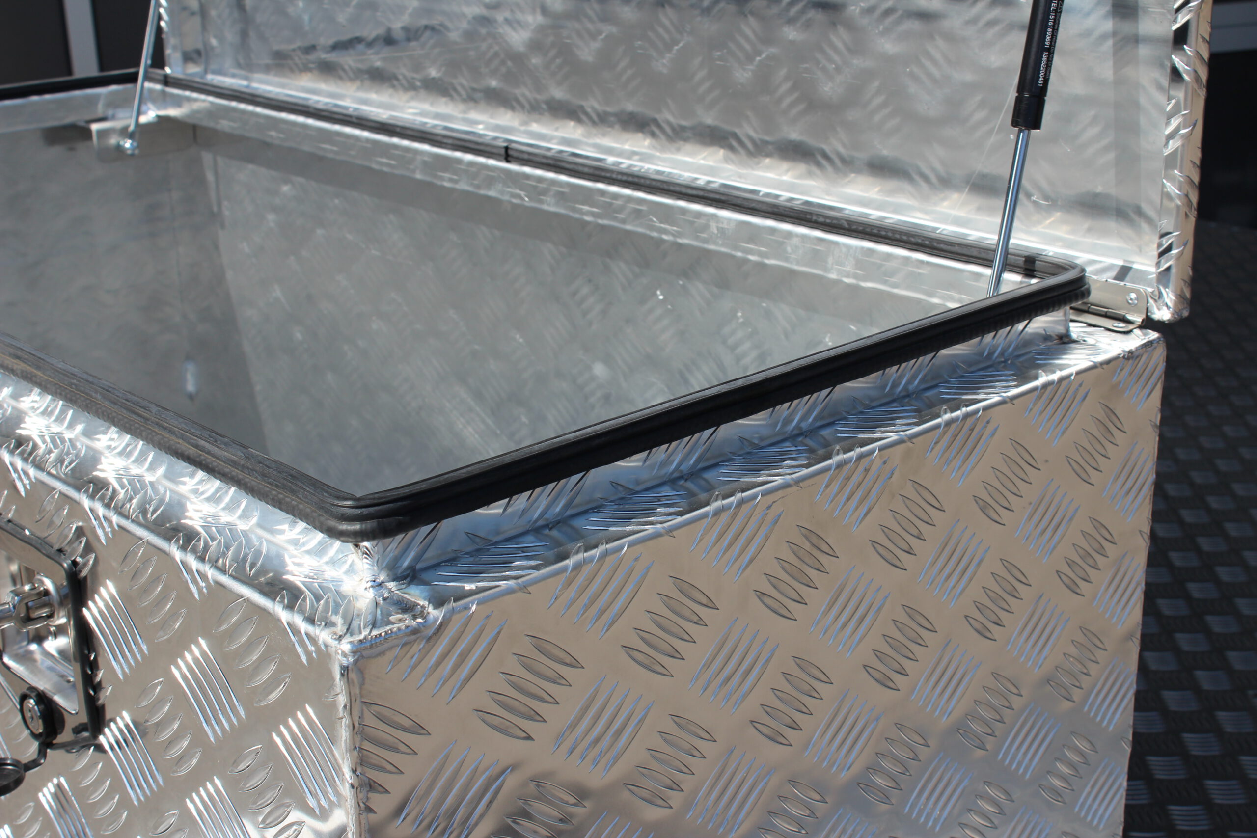 Große silberne Aluminium Riffelblech Staubox mit zwei Drehverschlüssen. Steht auf einem silbernen Aluminium Anhänger vor einem Tor