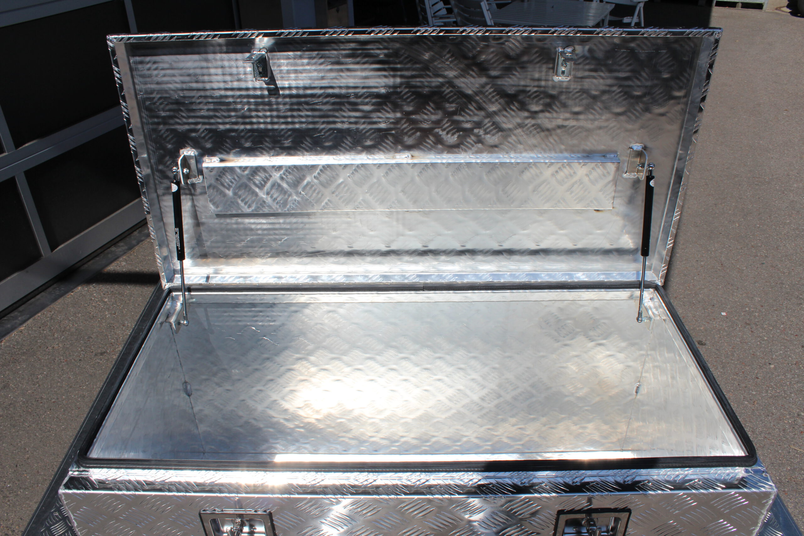 Große silberne Aluminium Riffelblech Staubox mit zwei Drehverschlüssen. Steht auf einem silbernen Aluminium Anhänger vor einem Tor
