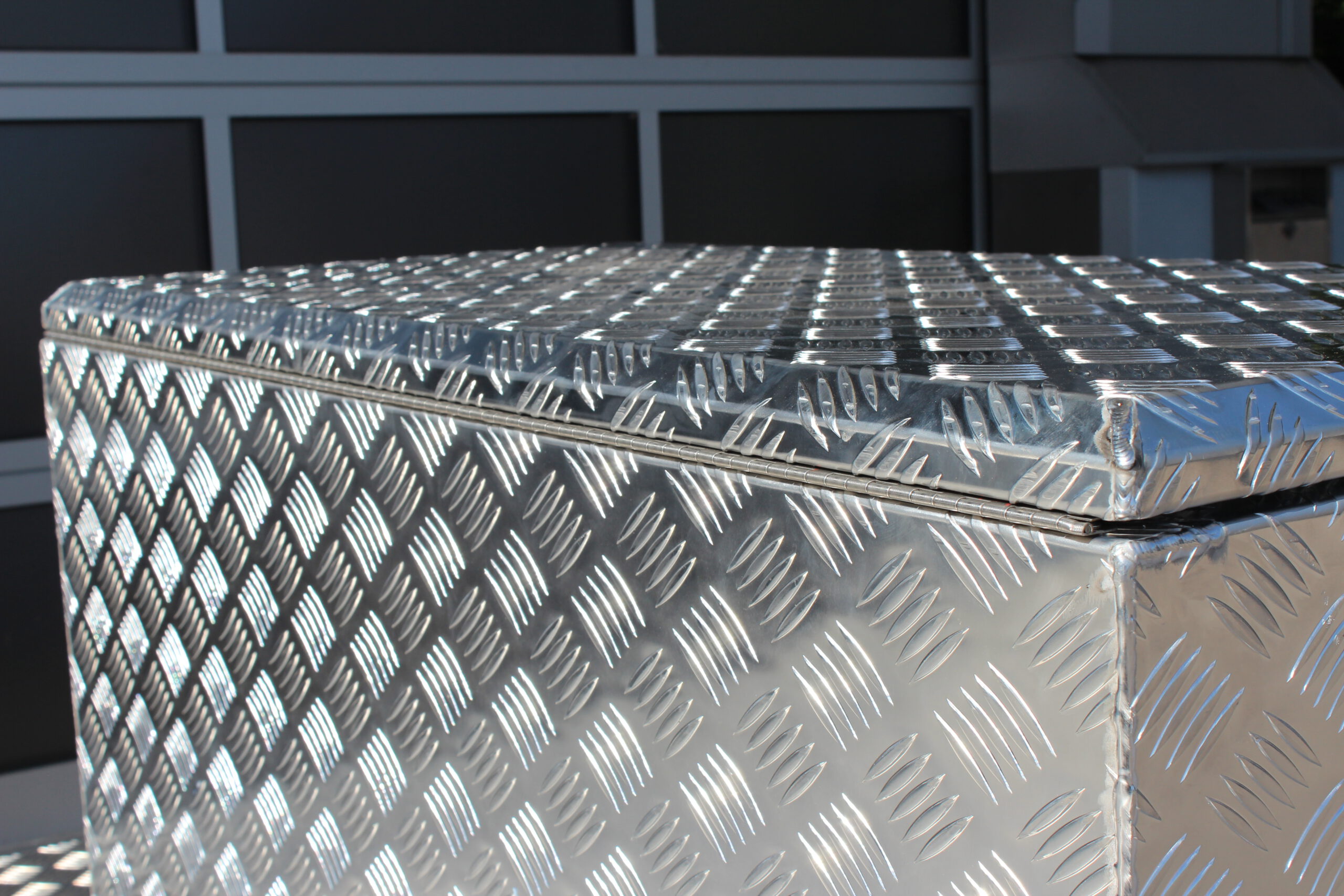 Große silberne Aluminium Riffelblech Staubox mit zwei Drehverschlüssen. Steht auf einem silbernen Aluminium Anhänger vor einem Tor